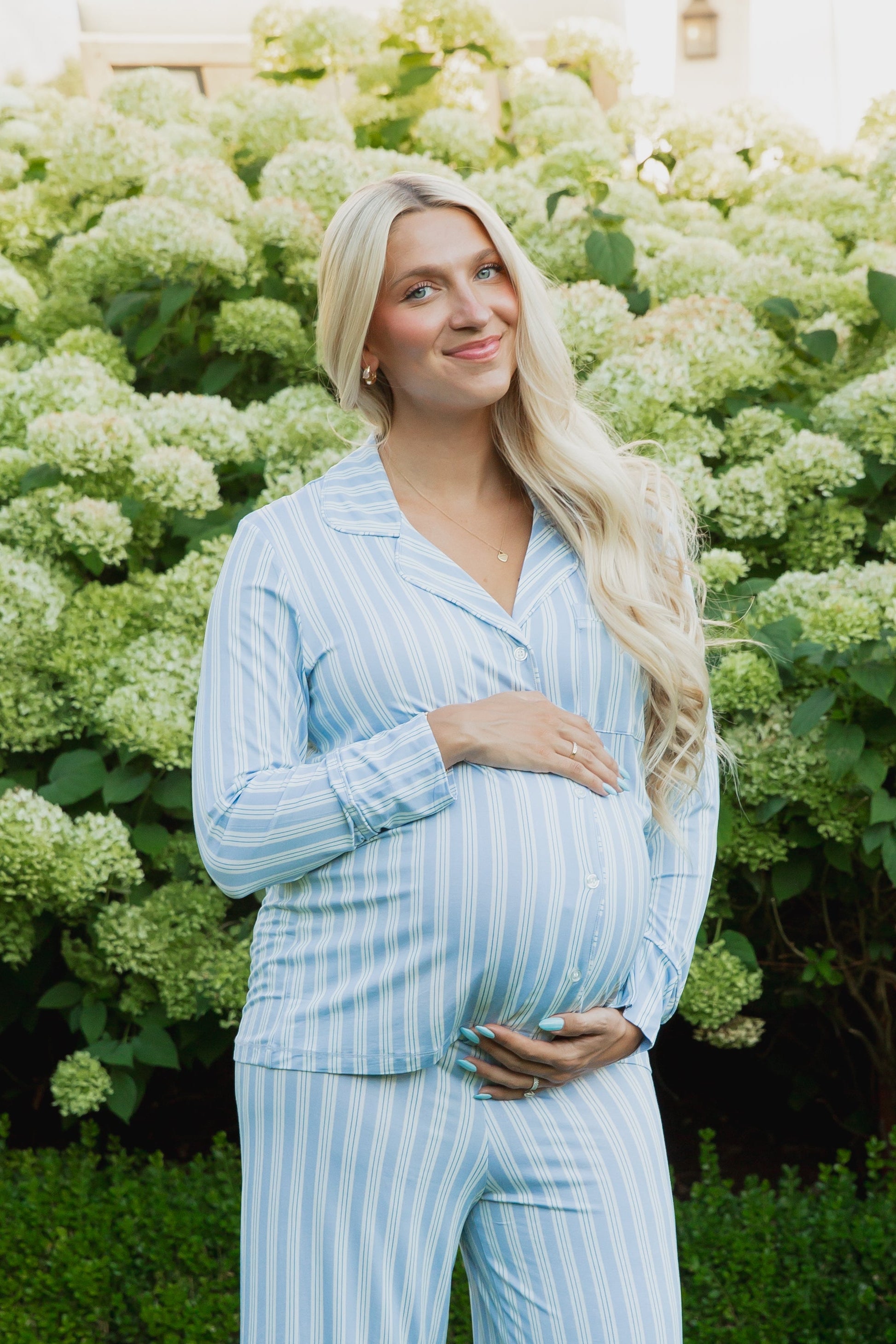 A pregnant woman with long blonde hair, wearing the haven pajama set with long pants from the Sarah Sweeten Goosebumps Collection in light blue stripes, stands outside in front of green hydrangea bushes, smiling and cradling her belly with both hands.