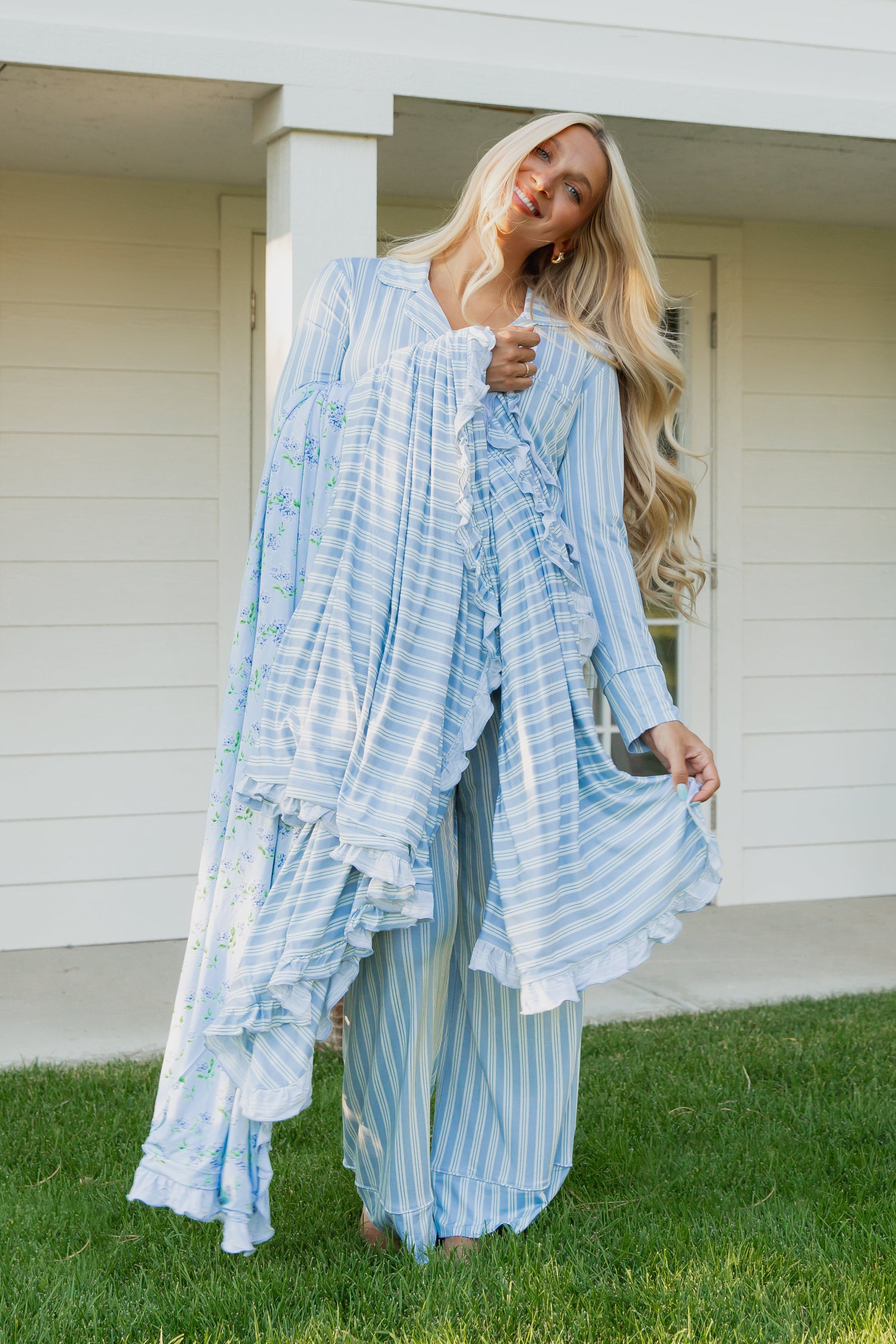 A woman with long blonde hair stands on the grass, smiling as she wears the haven / ruffle loose, light blue striped pajamas with ruffles. She holds part of her robe from the collection and has a luxurious blanket draped over her arm while posing in front of a white building with pillars.