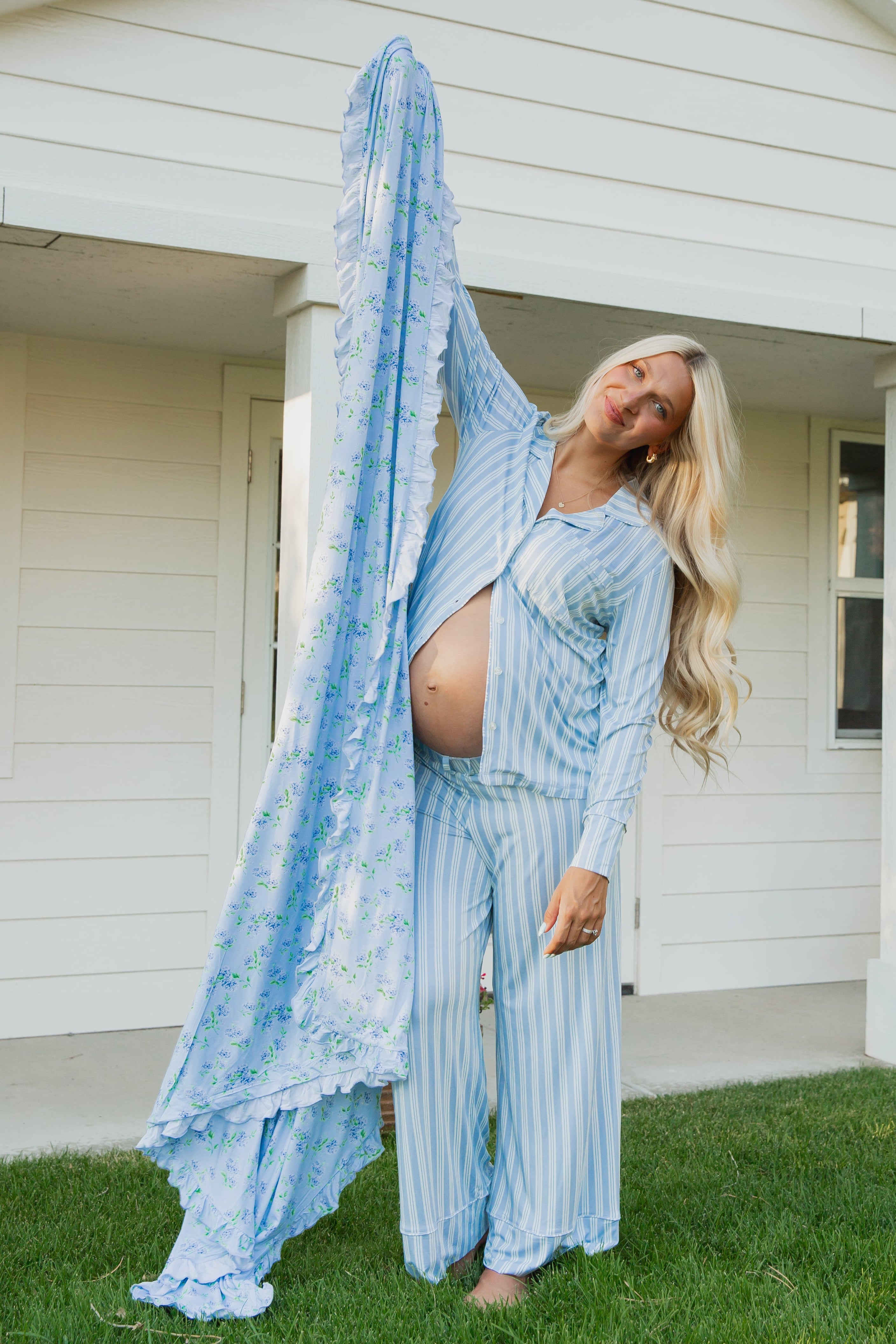 A pregnant woman in blue striped pajamas stands barefoot on the grass, smiling with her head tilted as she holds up the hydrangea / ruffle bamboo blanket in front of a white house.