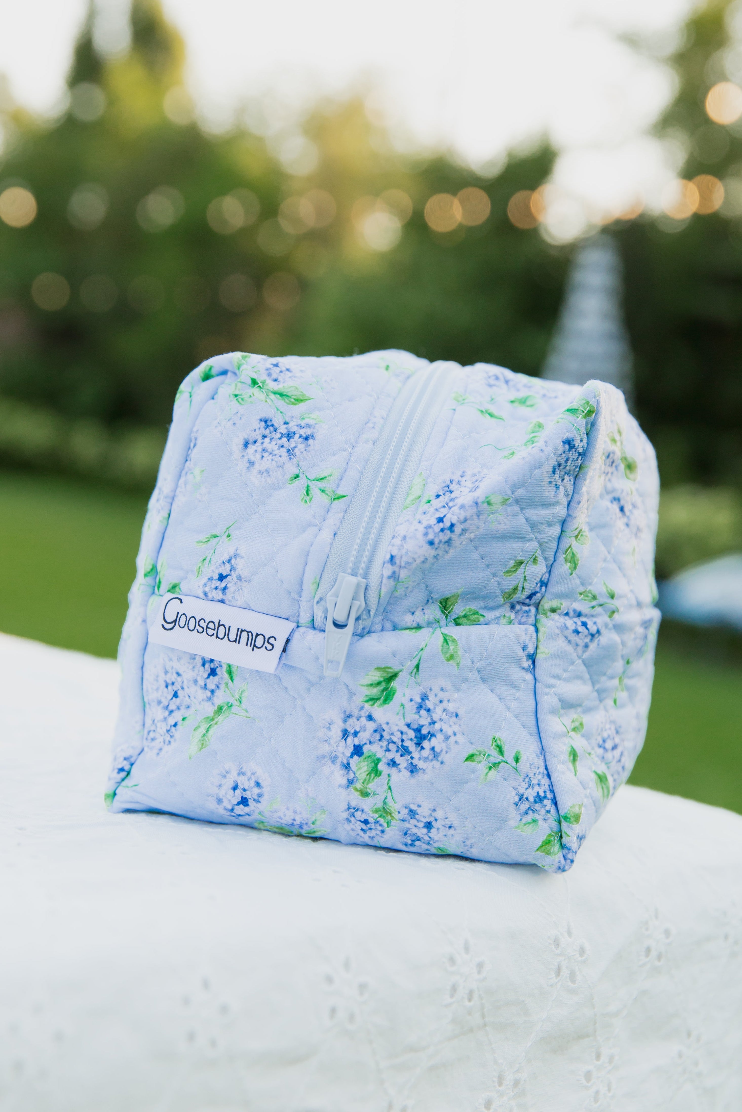 A soft, quilted, cube-shaped pouch with a delicate hydrangea print and a zipper sits on a white outdoor surface. A label reads “hydrangea / travel bag.”.