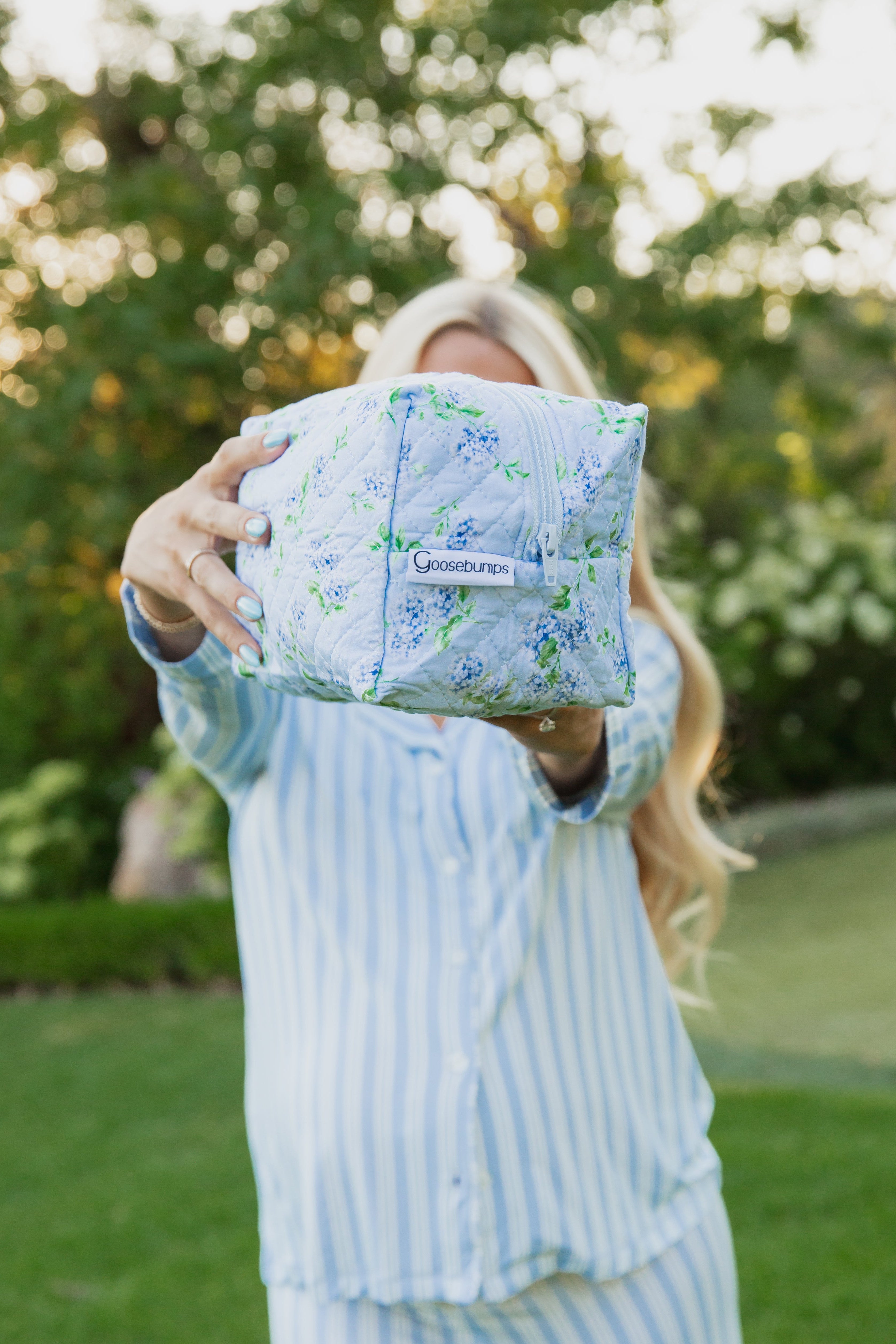 A person in blue striped pajamas holds the hydrangea / travel bag toward the camera, its floral hydrangea print obscuring their face. They stand outside with greenery and trees in the background, showcasing a piece from the Sarah Sweeten Collection.