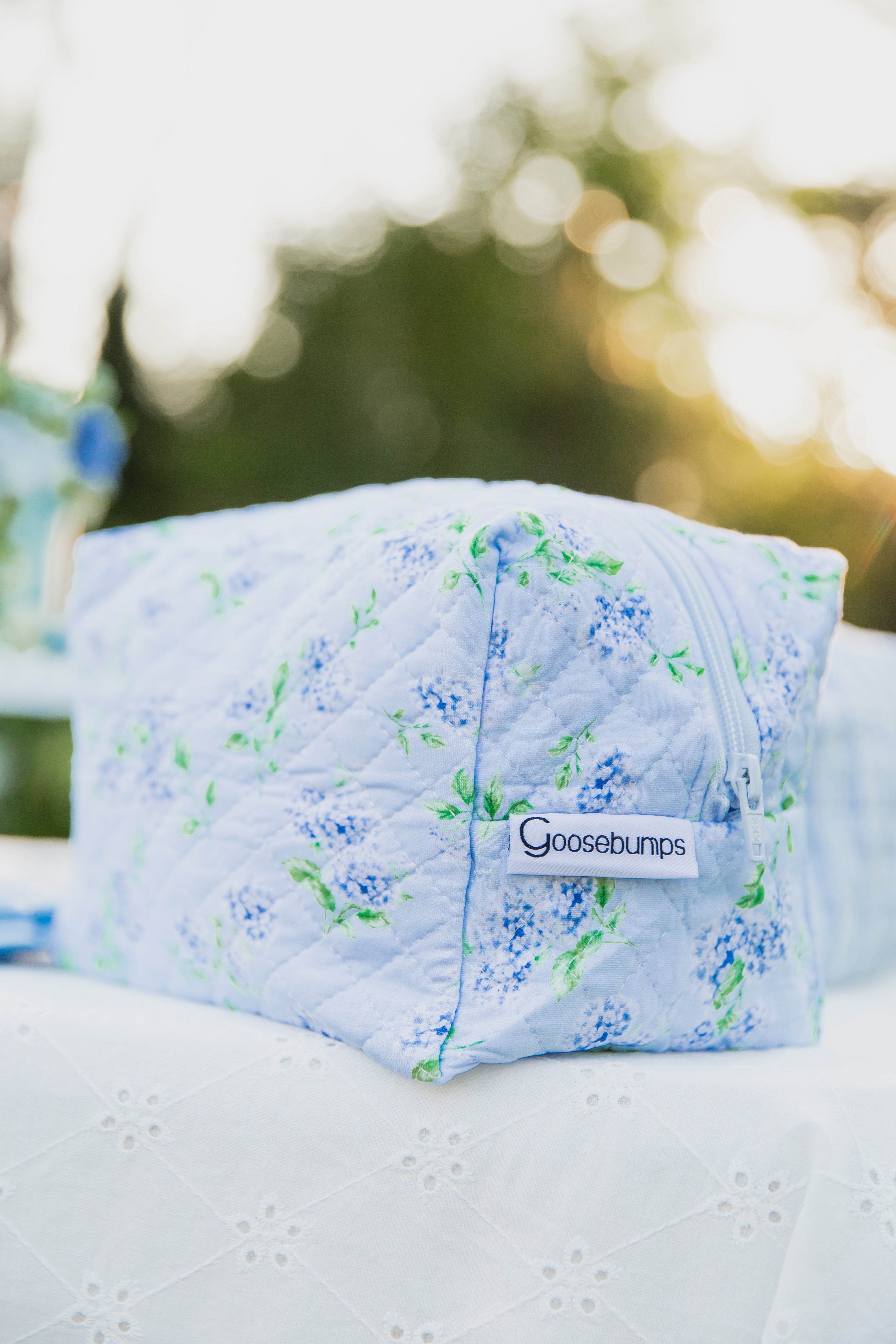 A quilted fabric pouch featuring a hydrangea print from the hydrangea / travel bag collection rests on a white embroidered cloth outdoors, surrounded by blurred greenery and sunlight in the background.