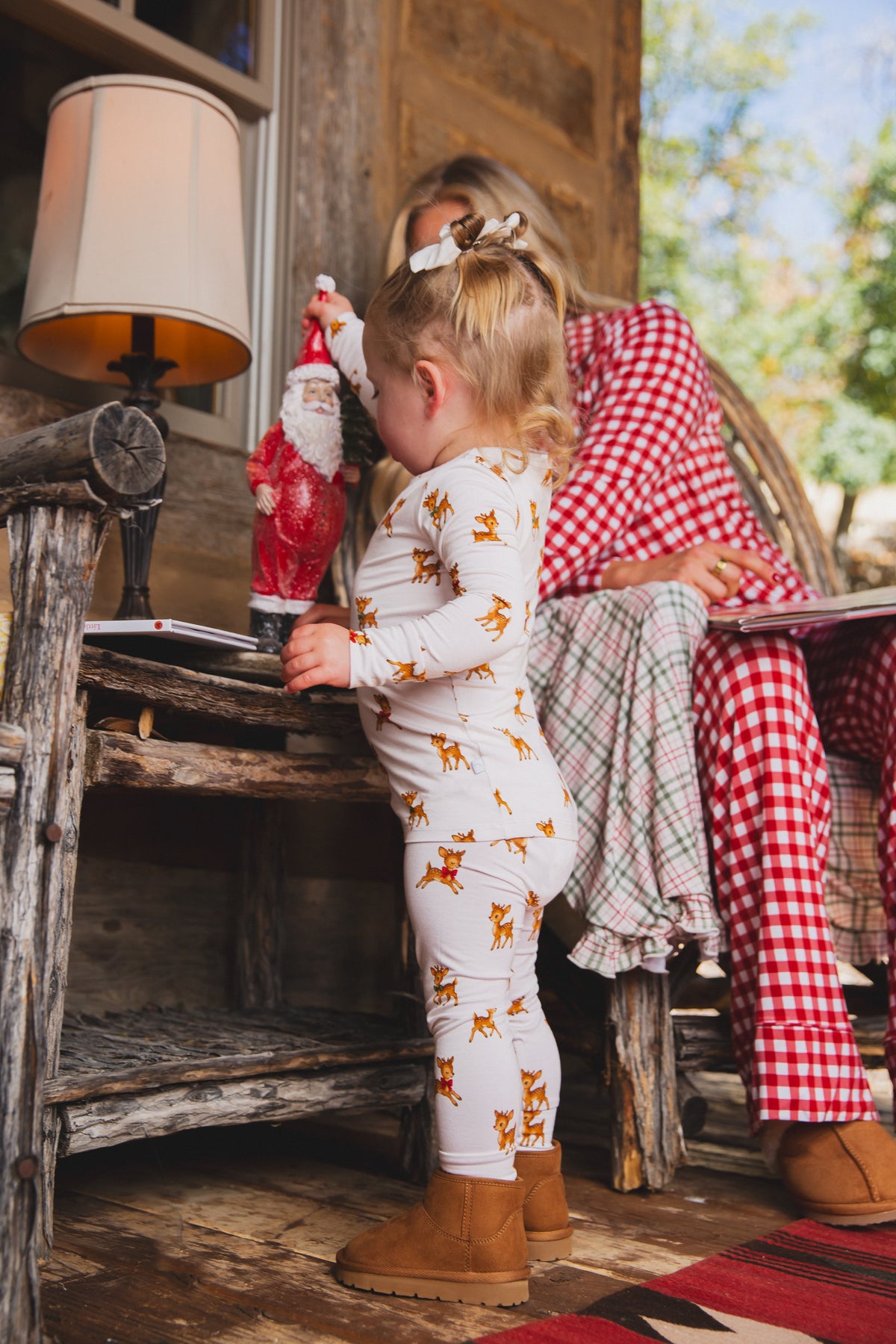 A toddler wears vintage reindeer / two piece skinny bamboo pajamas while standing by a wooden table and reaching for a Santa figurine.