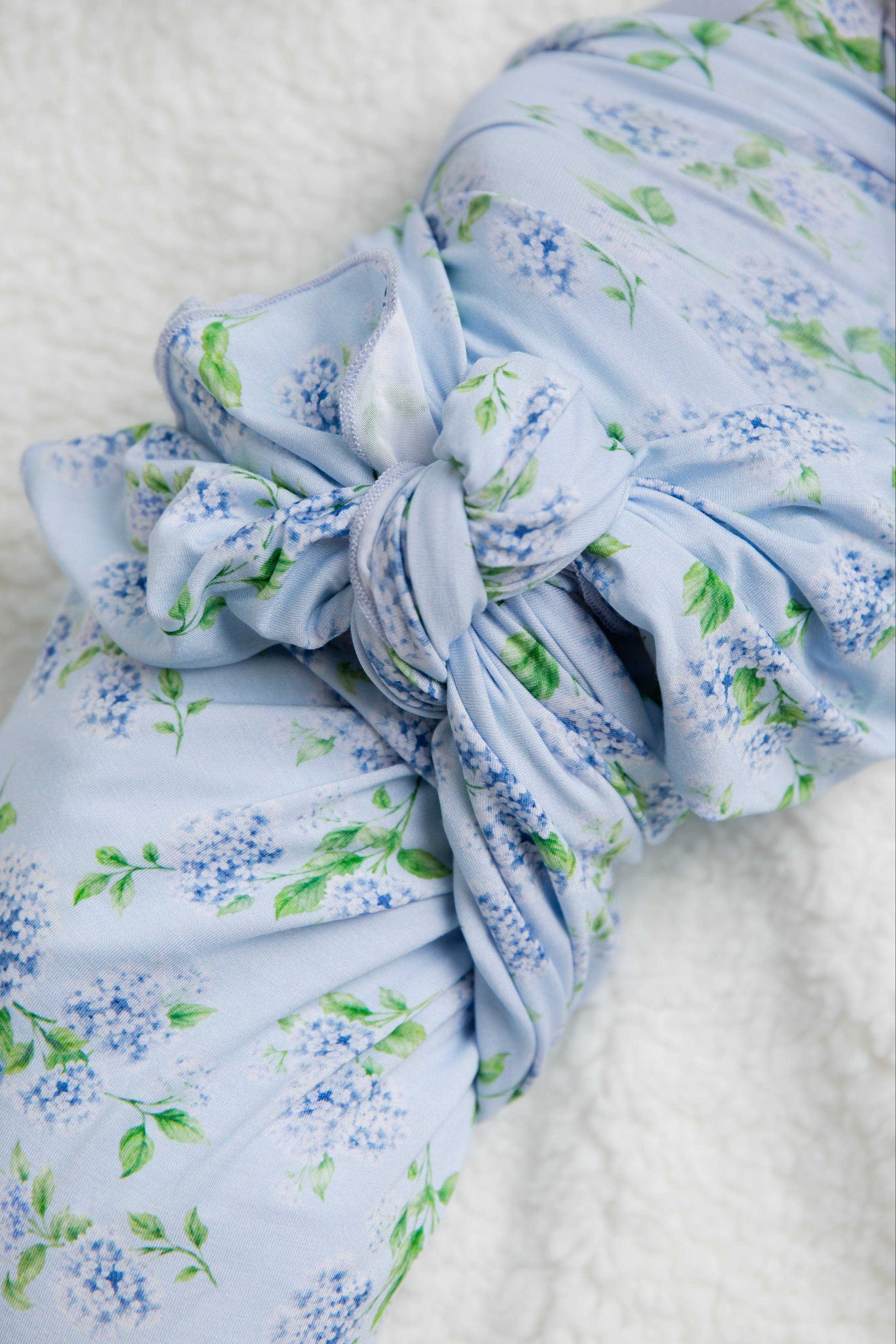 A close-up of a blue floral knot from the “hydrangea / swaddle,” part of the Sarah Sweeten Goosebumps Collection, showcases its delicate hydrangea print resting on a soft white textured surface.