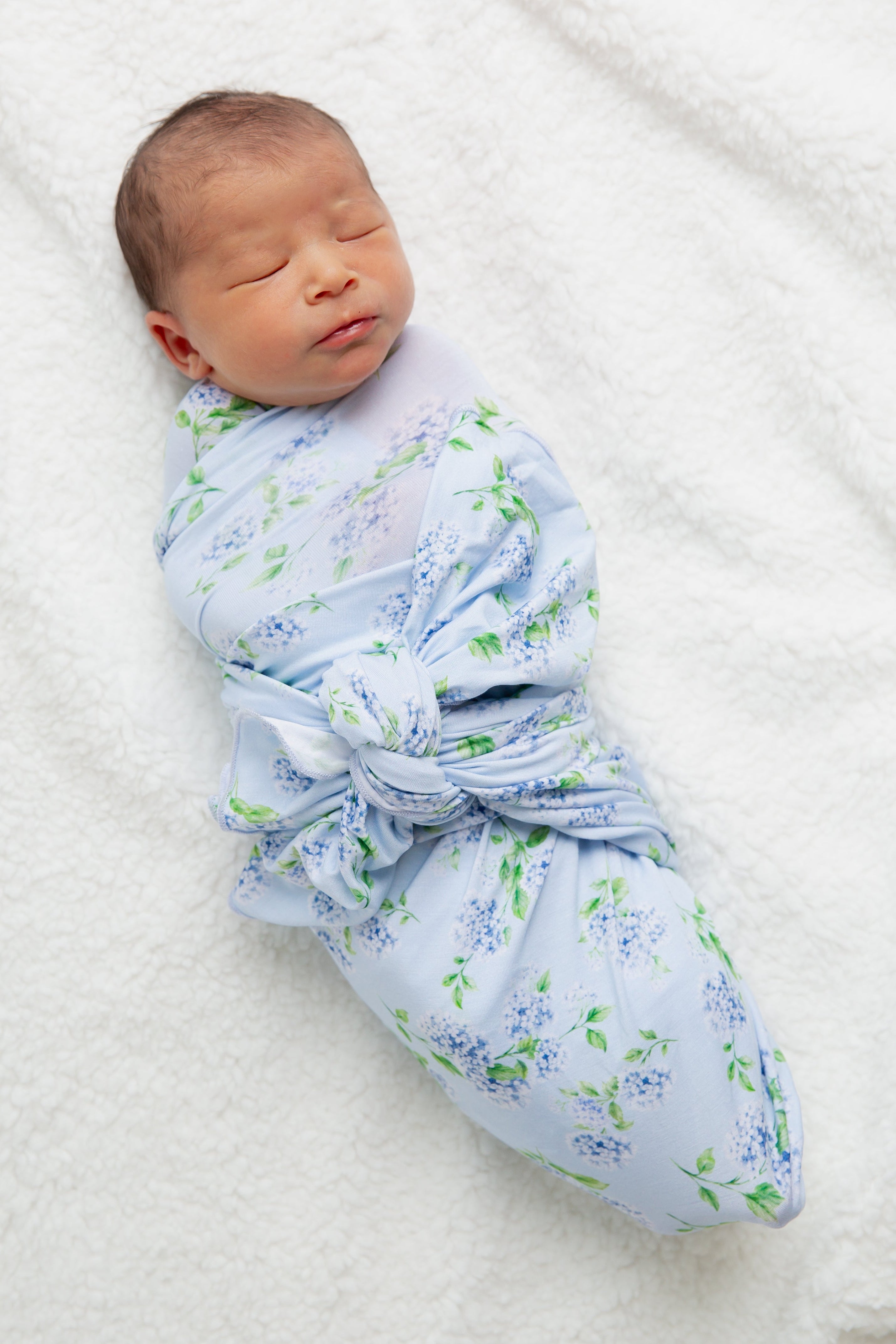 A newborn baby is peacefully swaddled in the hydrangea / swaddle, featuring a light blue hydrangea print from the Sarah Sweeten Goosebumps Collection, while lying on a soft white surface.