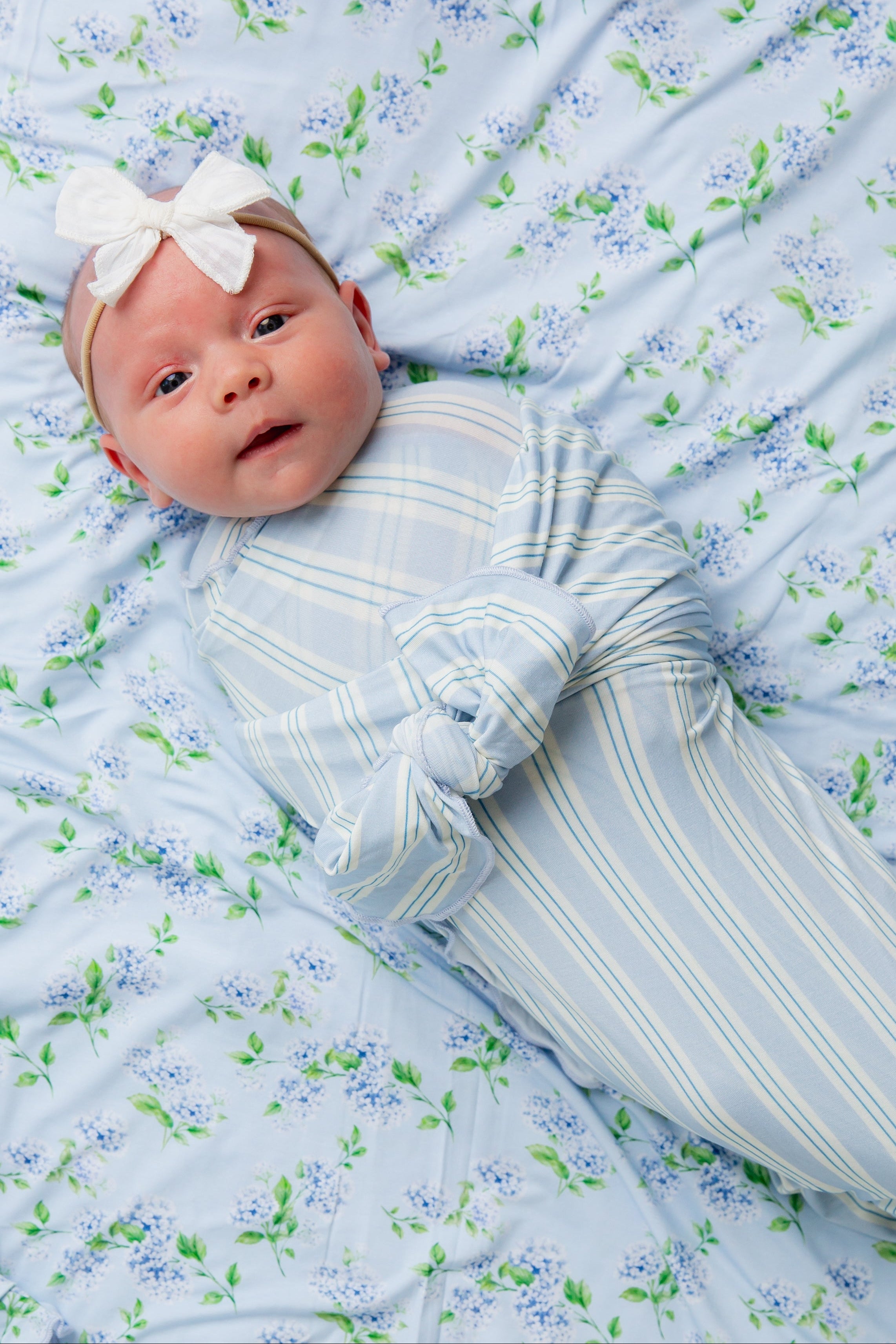 Wrapped in the haven / swaddle, featuring blue and white stripes from the Goosebumps Collection, a baby lies atop a blue floral blanket, wearing a white bow headband and gazing up with wide eyes.