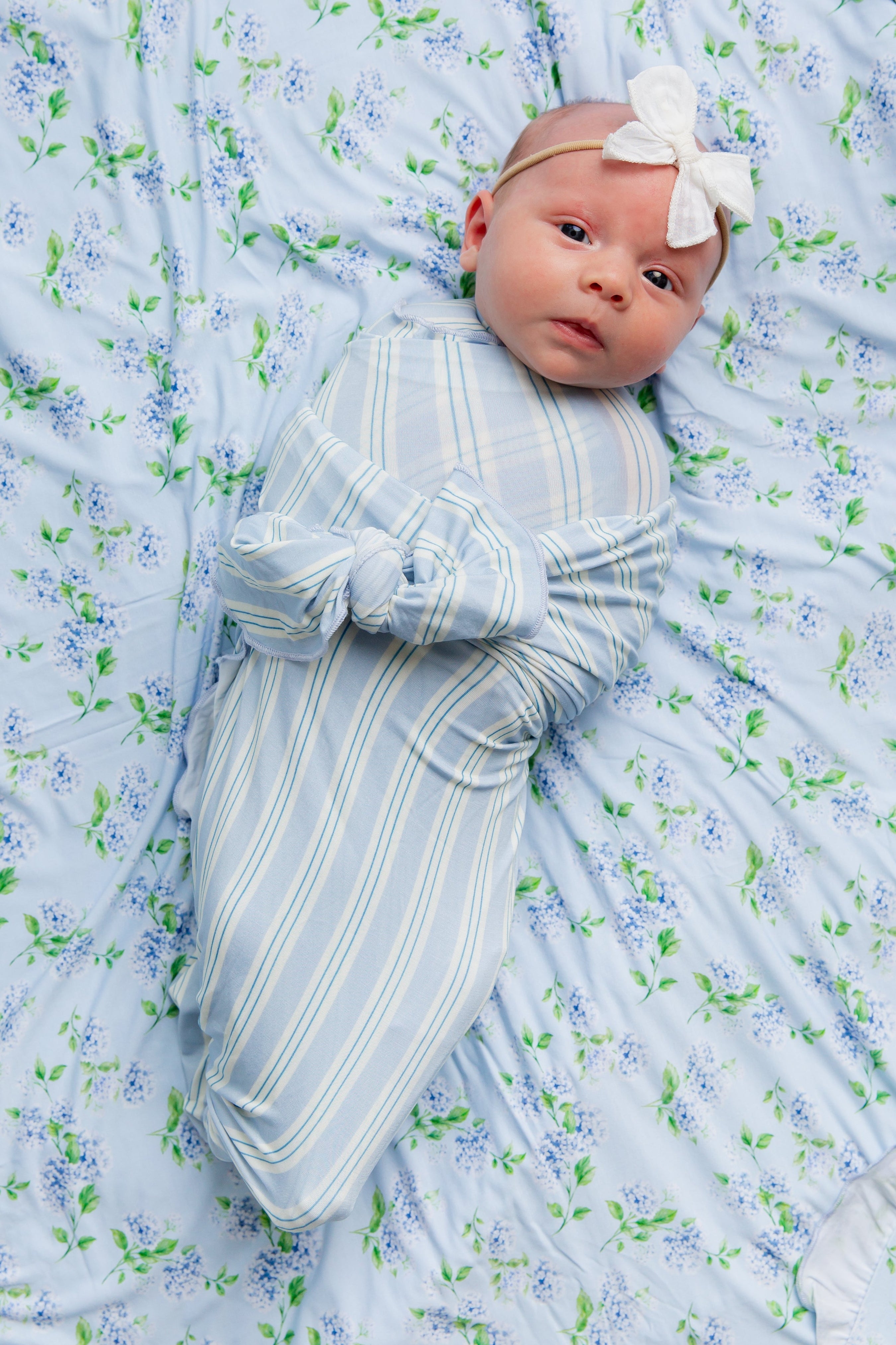 A baby wrapped in the haven / swaddle from the Goosebumps Collection, featuring blue and white stripes, lies on a light blue floral sheet while wearing a white bow headband and looking up.