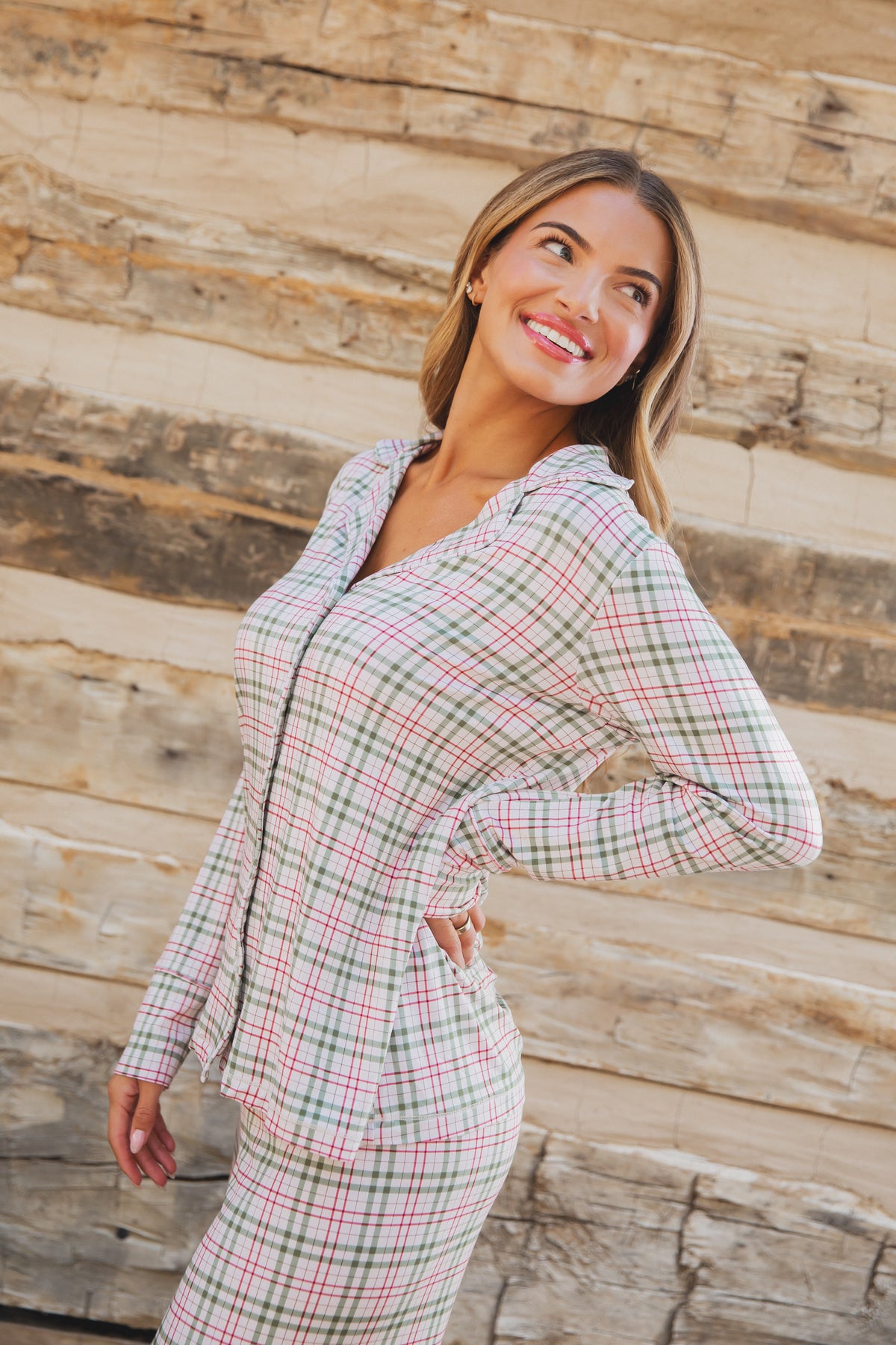 A smiling woman wears a red and green plaid homestead / pajama set / long pant long-sleeved bamboo top.