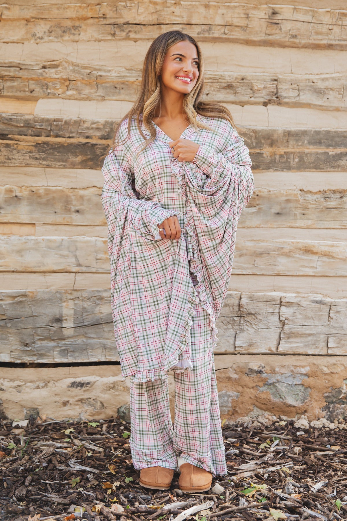 A woman wears a homestead / ruffle matching plaid pajama set with ruffle detailing and tan slippers while wrapped in a luxurious bamboo blanket in front of a log wall.