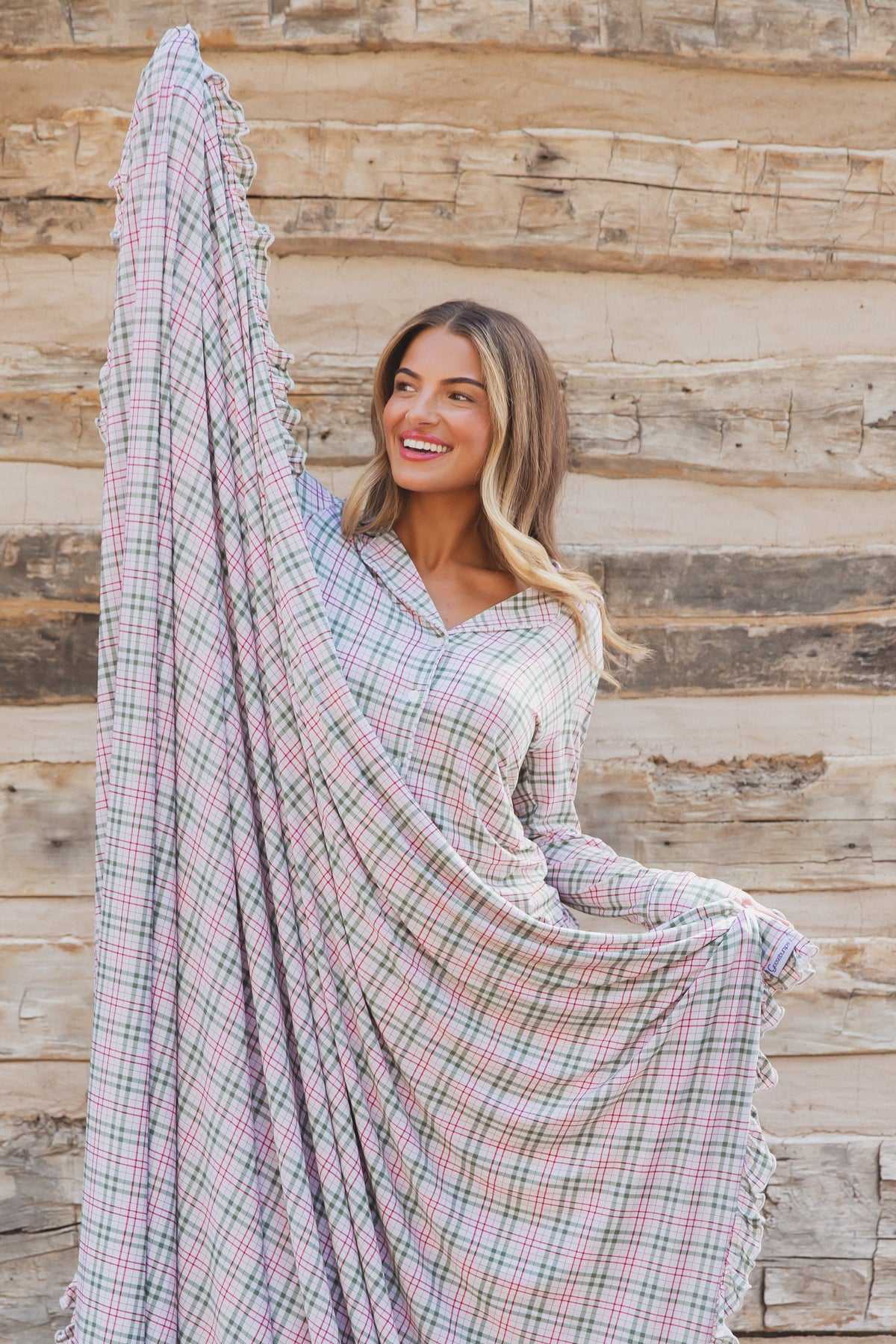 A smiling woman wearing a homestead / ruffle long-sleeved, green and red plaid nightgown with ruffle trim, holding up a section of the luxurious plaid blanket.