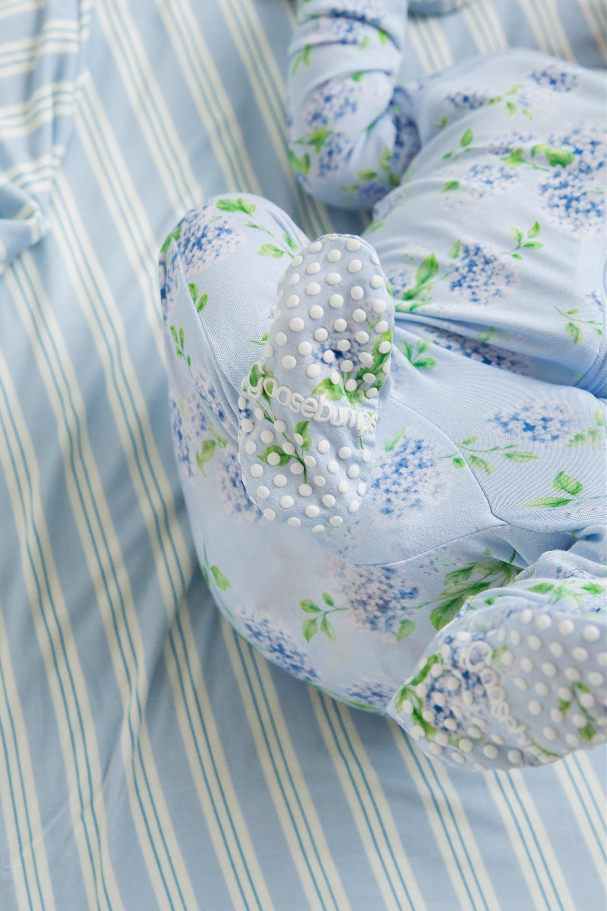 A baby’s legs in the hydrangea one piece zipper, featuring blue floral fabric with white grip dots on the feet, rest on a blue and white striped sheet, while the buttery-soft material ensures ultimate comfort for your little one.