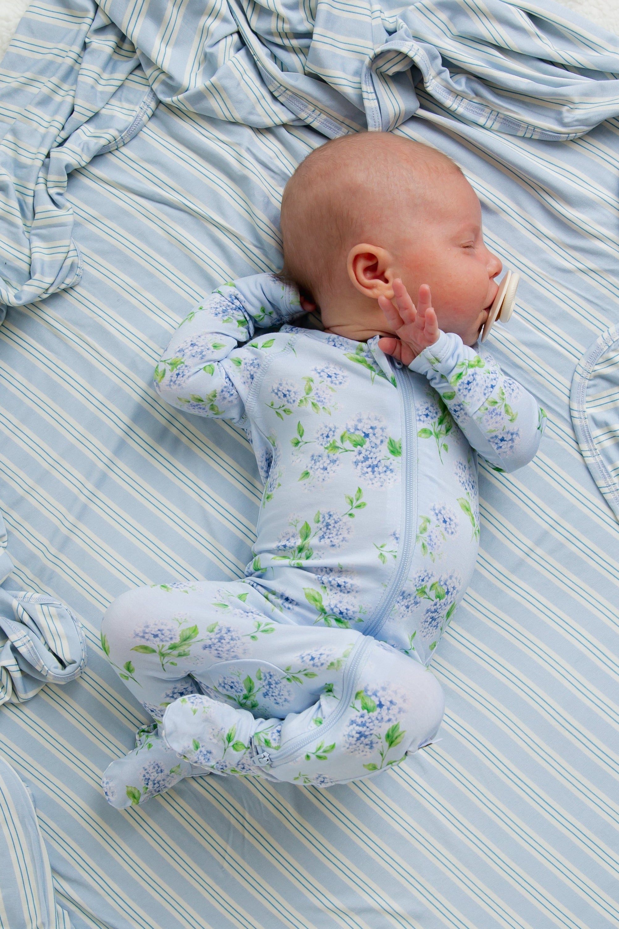 A baby wearing the hydrangea one piece zipper, crafted from buttery-soft fabric in a blue floral pattern, lies asleep on a blue and white striped blanket with a pacifier in their mouth and one hand near their face.
