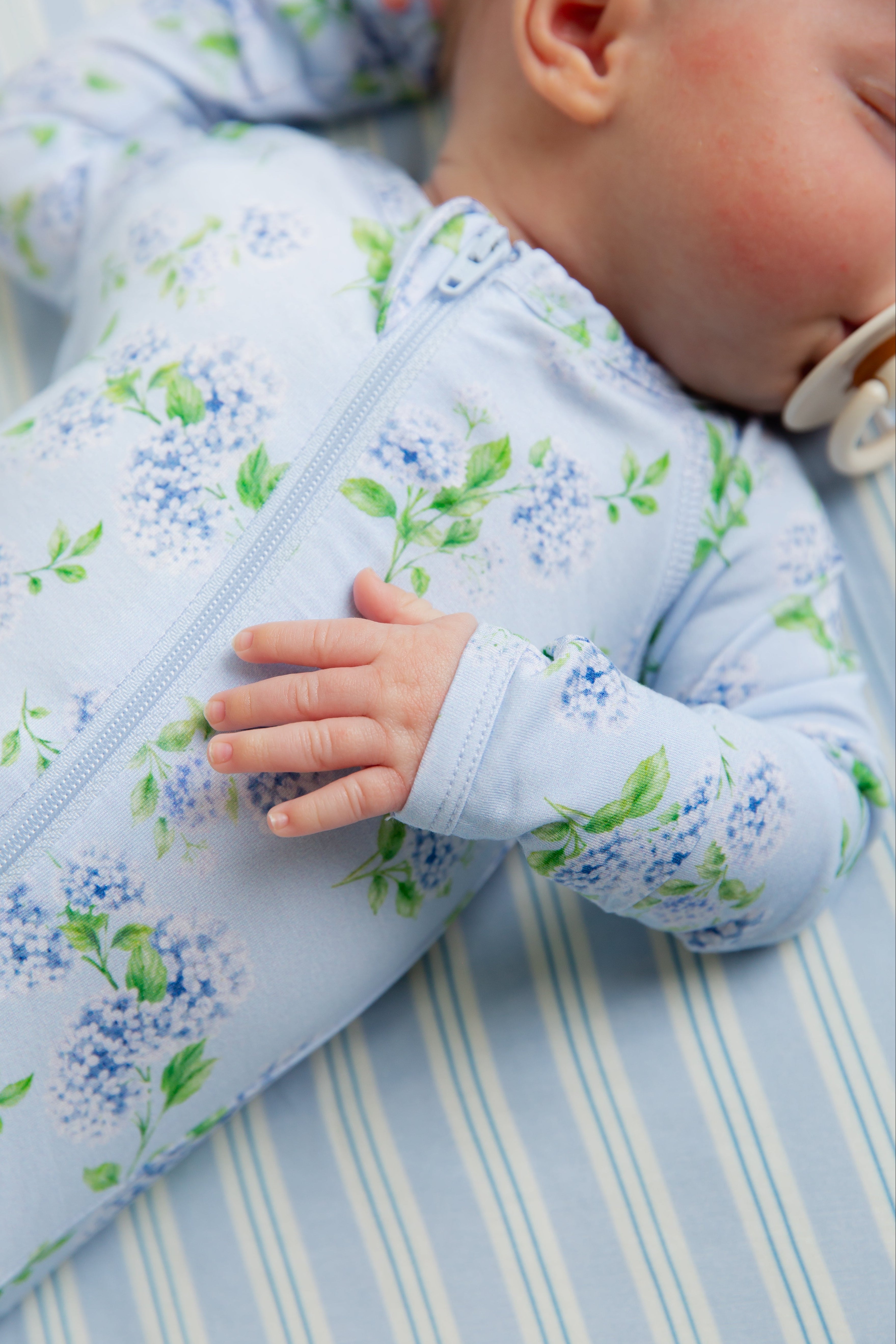 A baby wearing the hydrangea one piece zipper, made from buttery-soft fabric, sleeps on a blue and white striped sheet with a pacifier in their mouth and one hand resting on their chest.