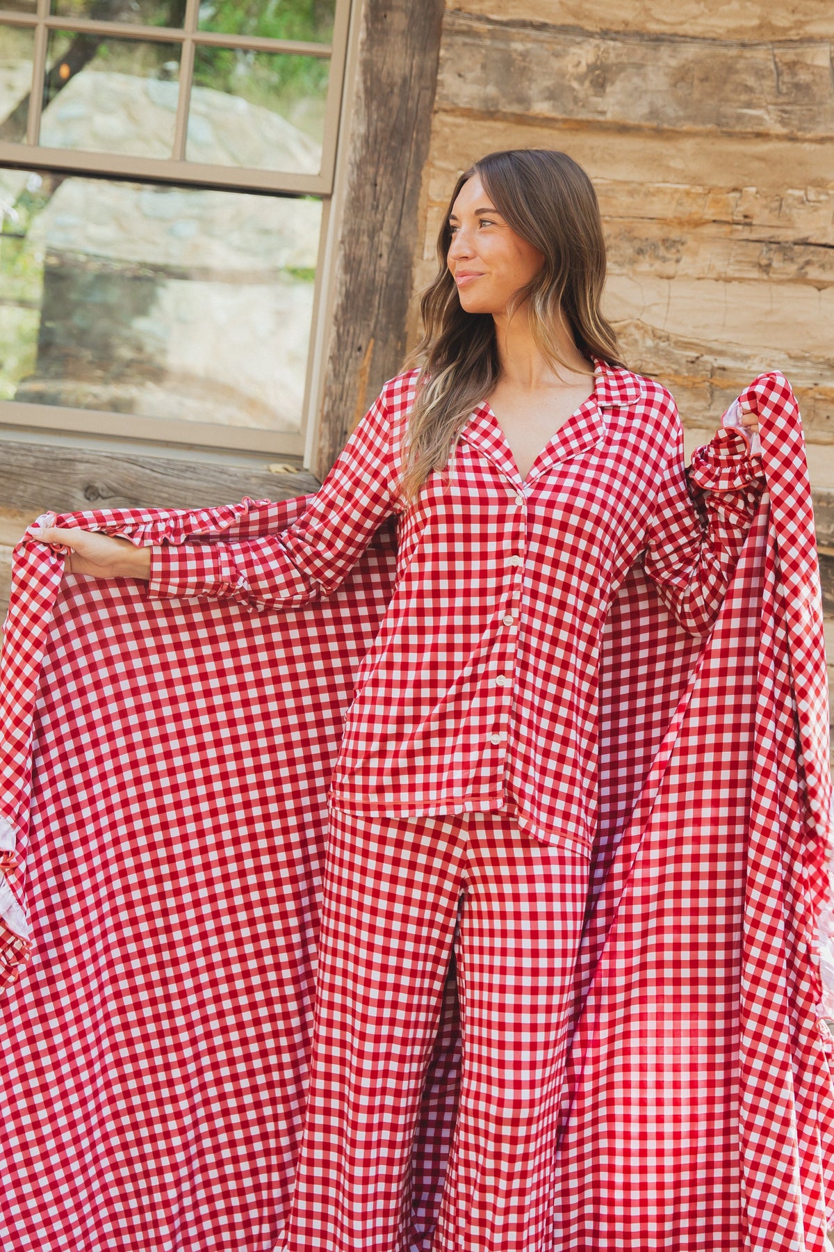 A woman wears the checkin’ it twice / ruffle pajama set in red and white gingham with ruffled cuffs and matching pants, while holding out a large matching gingham blanket.