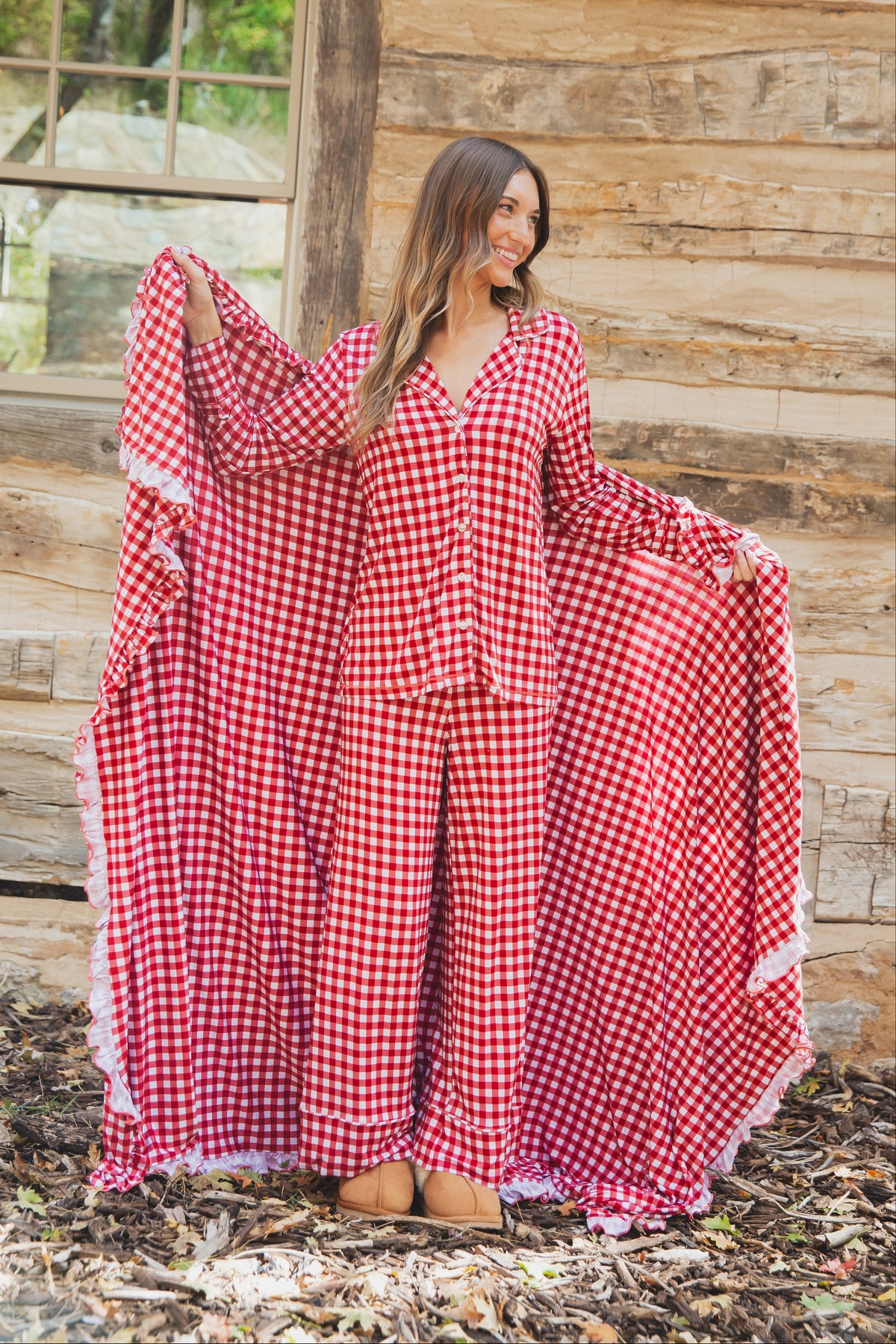 A woman stands outdoors wearing checkin’ it twice / ruffle red and white gingham pajamas with ruffles while holding up a matching ruffle-trimmed bamboo blanket.