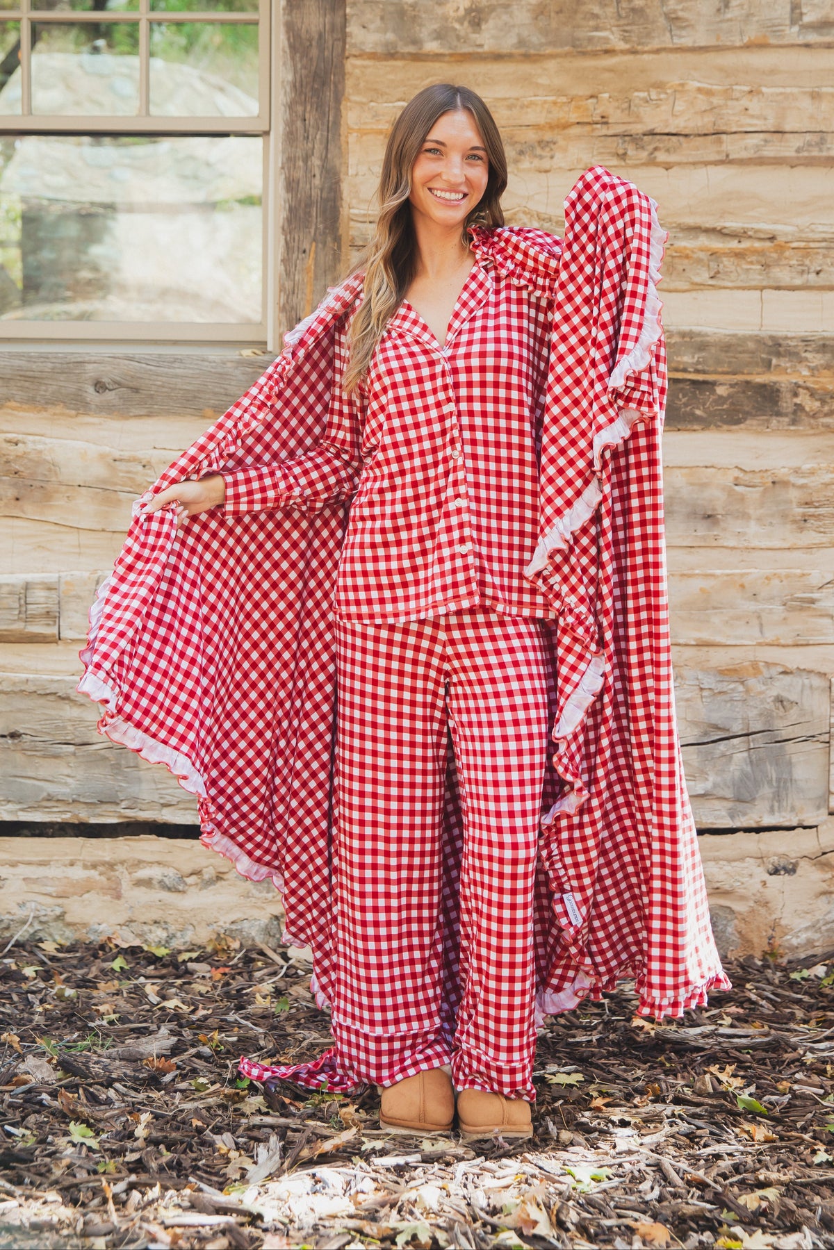 A smiling woman wears the red and white checkin’ it twice / ruffle button-down pajama set with ruffled trim, holding out a coordinating, luxurious bamboo blanket.