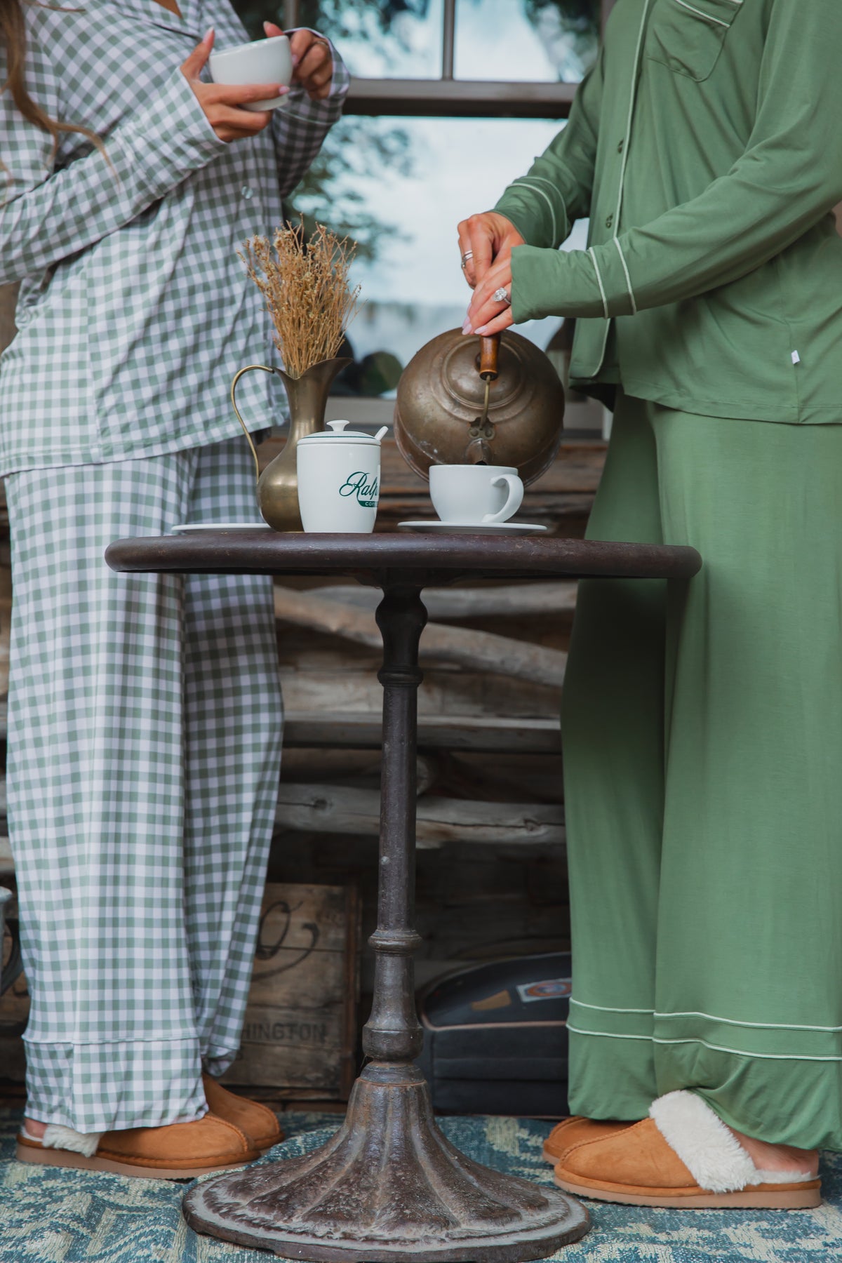 Two people wearing evergreen lane / pajama set / long pant in green gingham stand at a small bistro table, one pouring tea from a metal kettle into a cup.