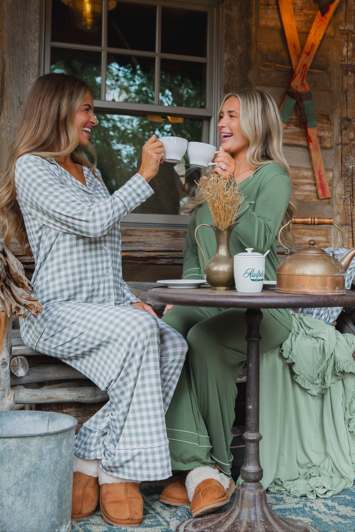 Two women wearing evergreen lane / pajama set / long pant in green gingham sit at an outdoor table, smiling and clinking white coffee mugs together.