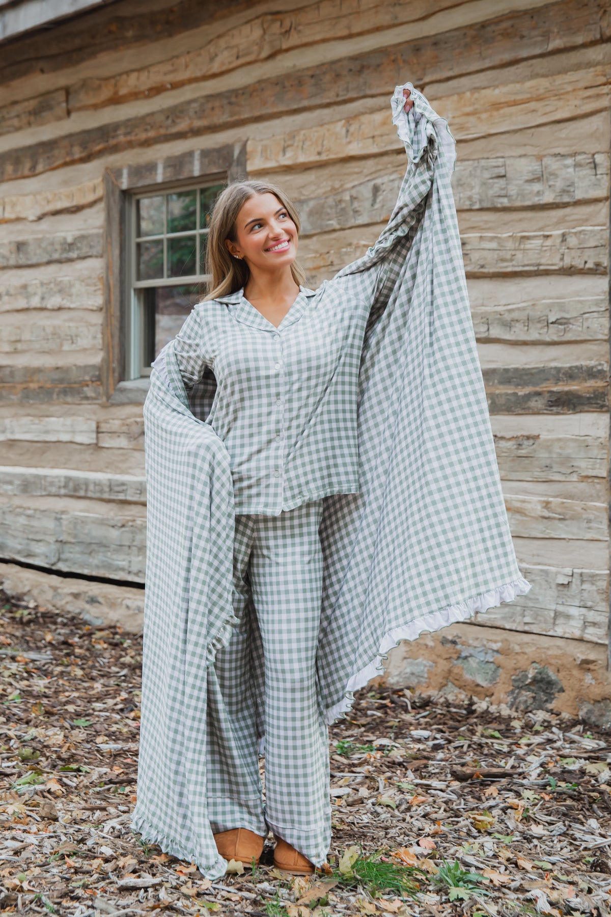 A person smiles while wearing an evergreen lane / ruffle pajama set with green and white gingham and ruffled edges, holding out a matching green gingham blanket.