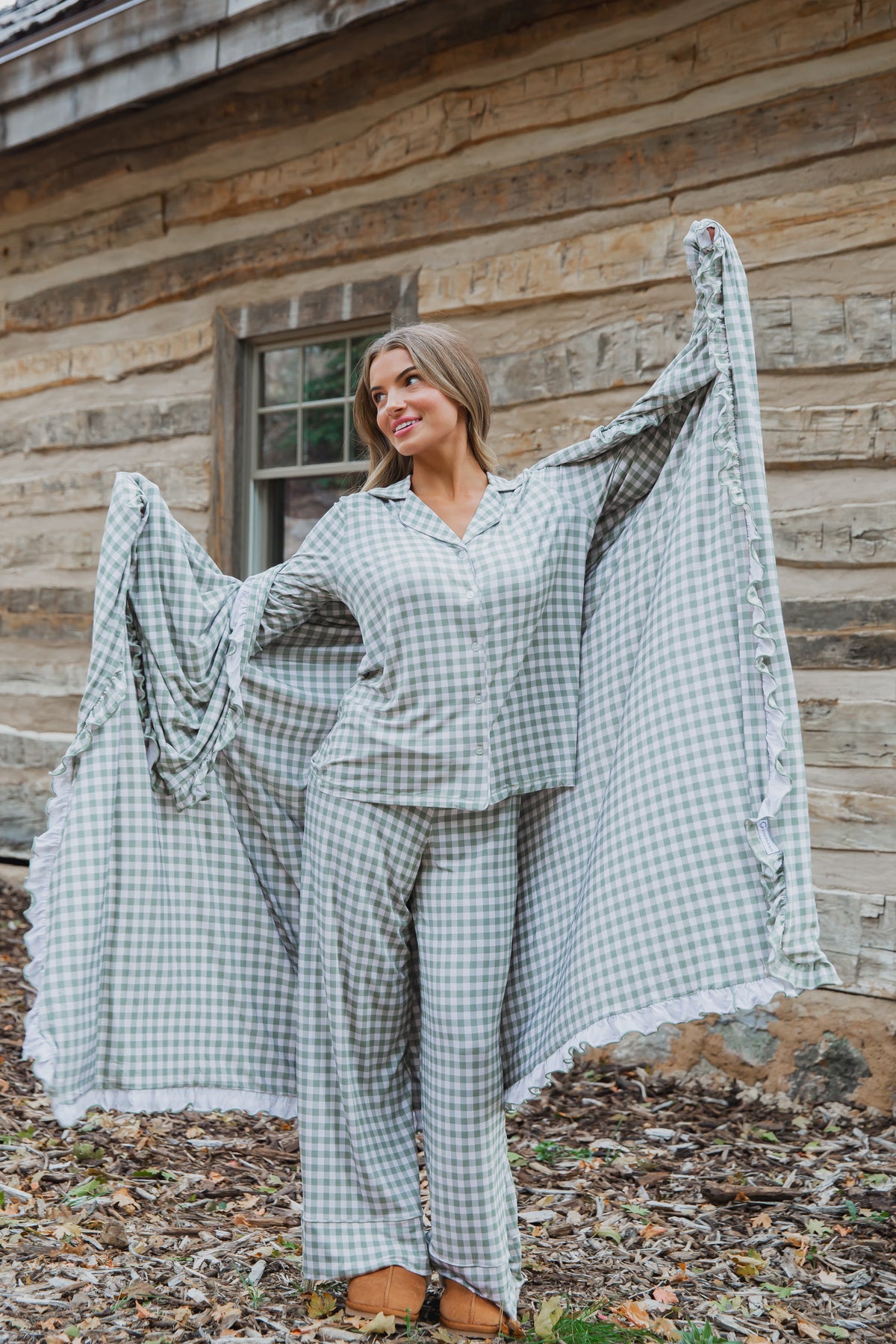 A woman wears an evergreen lane / ruffle green and white gingham button-down pajama set with ruffled trim while holding a matching bamboo ruffle blanket in front of a log cabin.