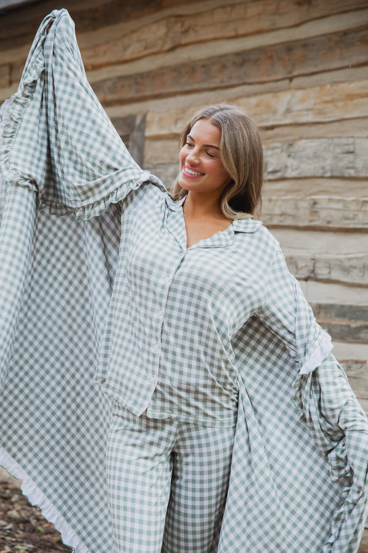 A smiling woman wears an evergreen lane / ruffle green and white gingham button-down pajama set with ruffled sleeves, coordinating with a matching ruffle blanket.