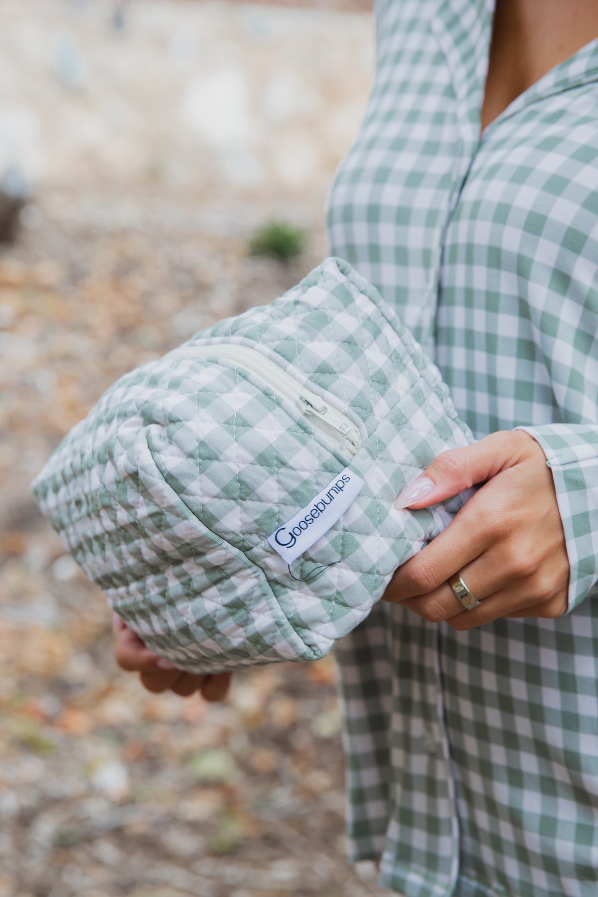 A person holds an evergreen lane / travel bag in a green and white gingham quilted fabric with a zippered closure.
