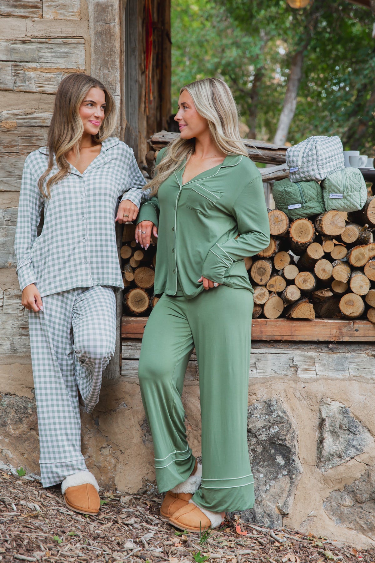 Two women stand outdoors wearing winter pine / pajama set / long pant, one in a green and white gingham set and the other in solid green with white piping, both wearing tan slippers.