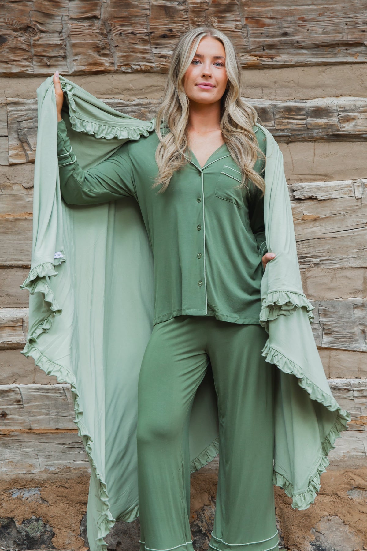 A woman wearing a frosted fir / ruffle pajama set with white piping, holding a matching ruffled bamboo blanket behind her.