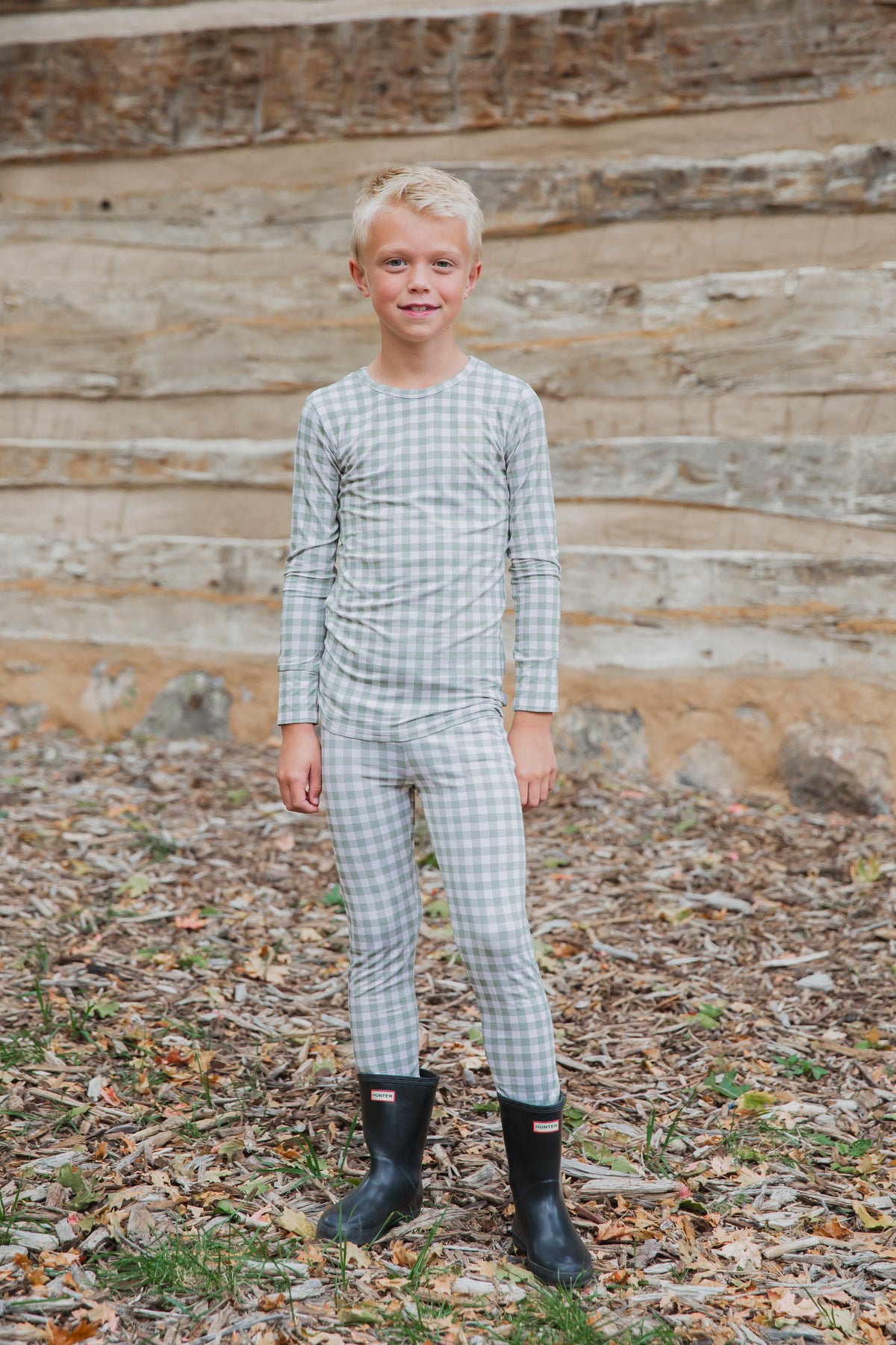 A young boy wearing an evergreen lane / two piece skinny in a sage green and white gingham print, styled as a long-sleeved shirt and matching leggings with black rubber boots.