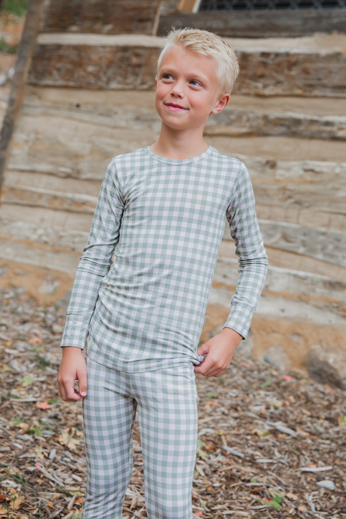 A young child smiles and looks off to the side while standing outdoors in evergreen lane / two piece skinny kids sleepwear, set against rustic wood siding with leaves and dirt on the ground.