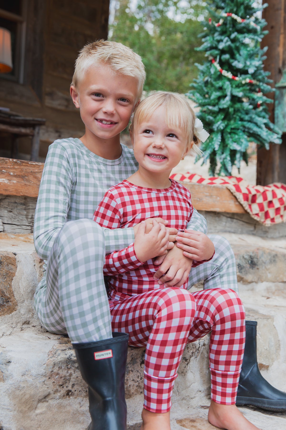 A boy and a girl wearing matching green and ivory gingham evergreen lane / two piece skinny bamboo pajamas sit on stone steps.