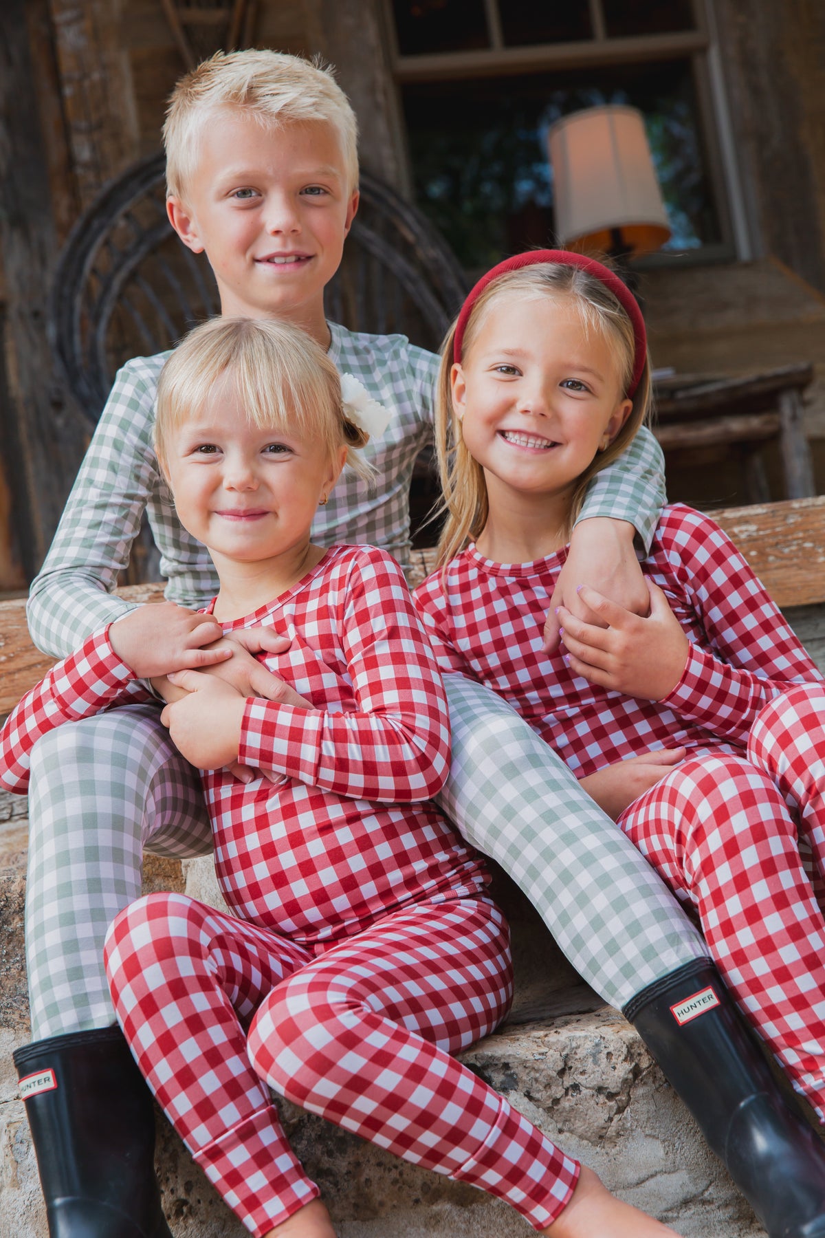 Three children sit on stone steps wearing evergreen lane / two piece skinny bamboo pajamas in red and green gingham patterns.