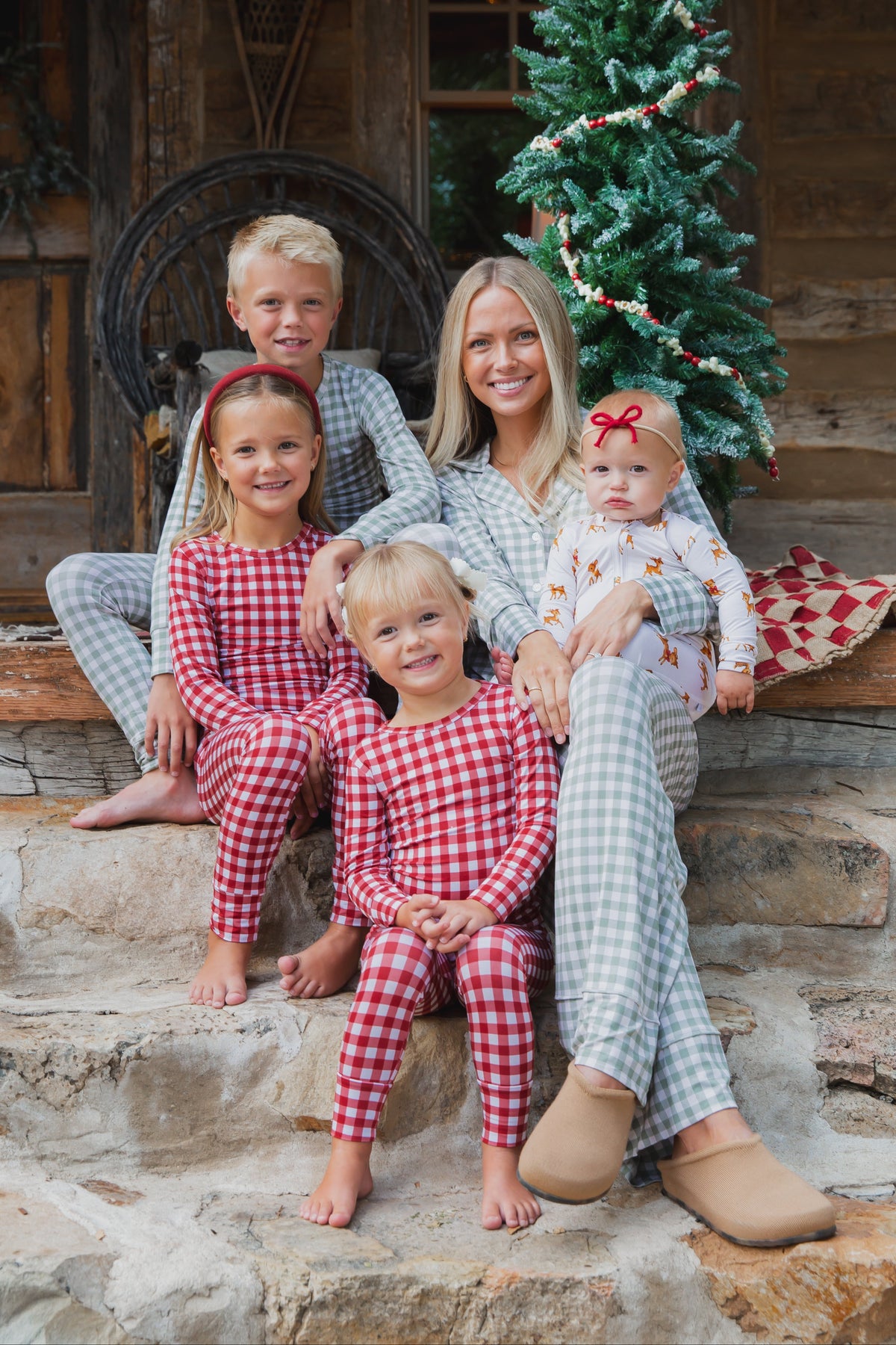 A woman and four children wearing red and white gingham checkin’ it twice / two piece skinny holiday pajamas sit on stone steps in front of a log cabin.