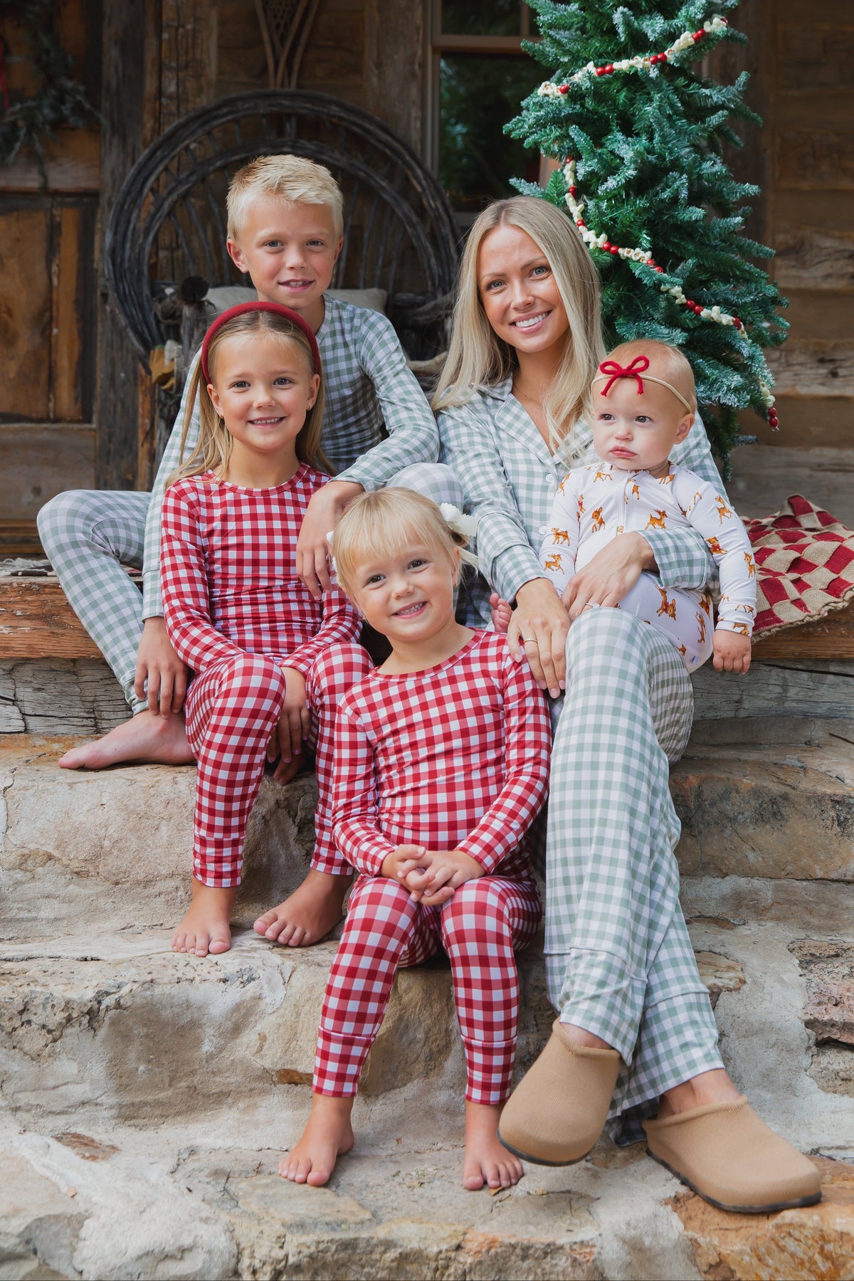 A woman and four children sit on stone steps in front of a Christmas tree wearing matching gingham pajamas, with one child in a vintage reindeer / one piece zipper newborn reindeer sleeper.
