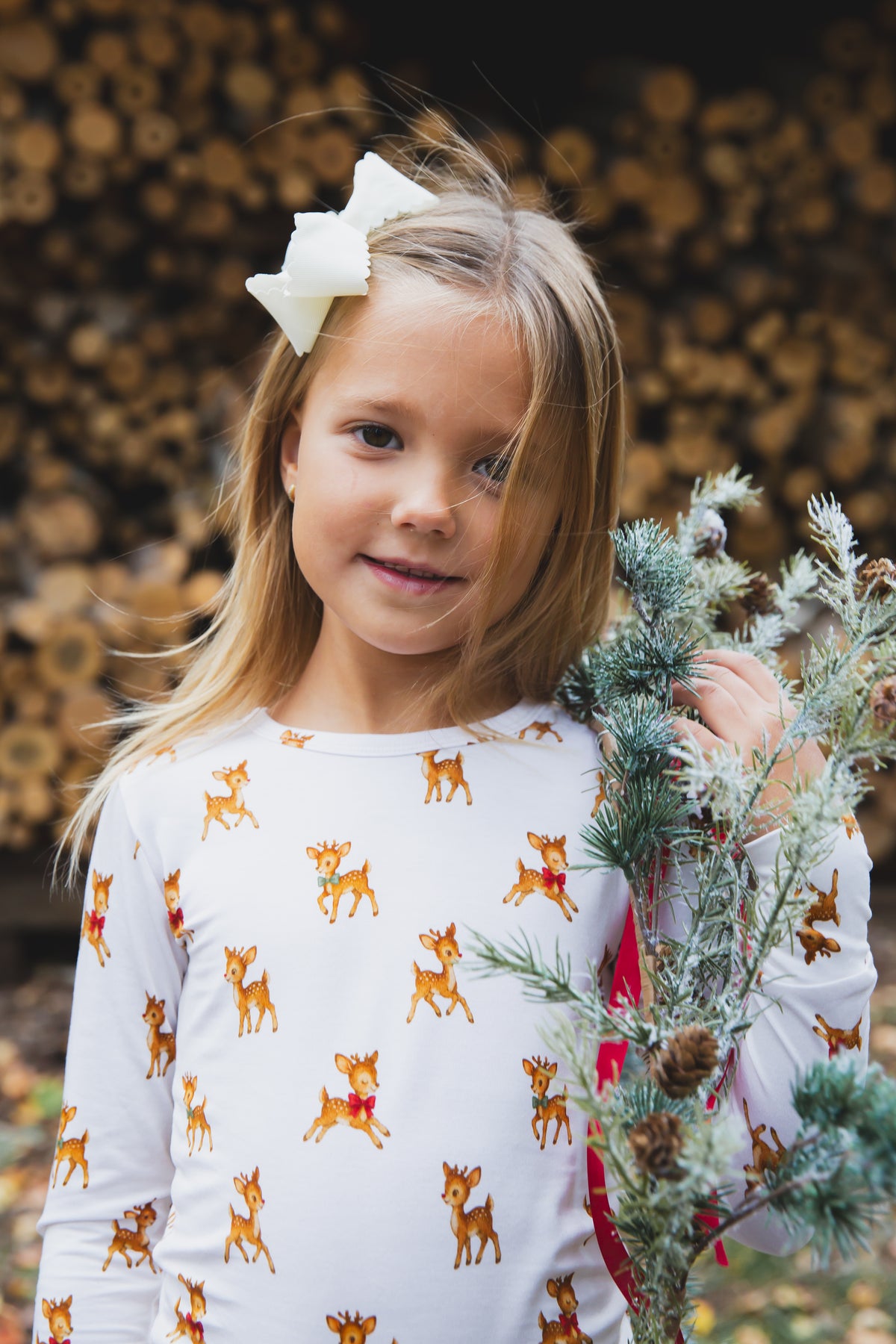 A girl with a white hair bow wears long-sleeved vintage reindeer / two piece skinny pajamas, holding a small artificial pine branch.