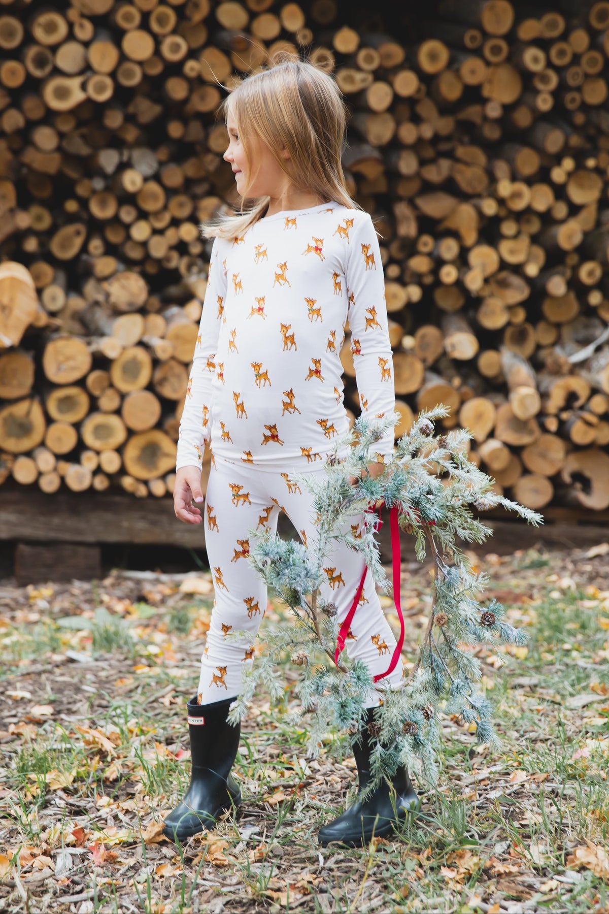 A child in vintage reindeer / two piece skinny bamboo pajamas and black rain boots, holding a holiday wreath.