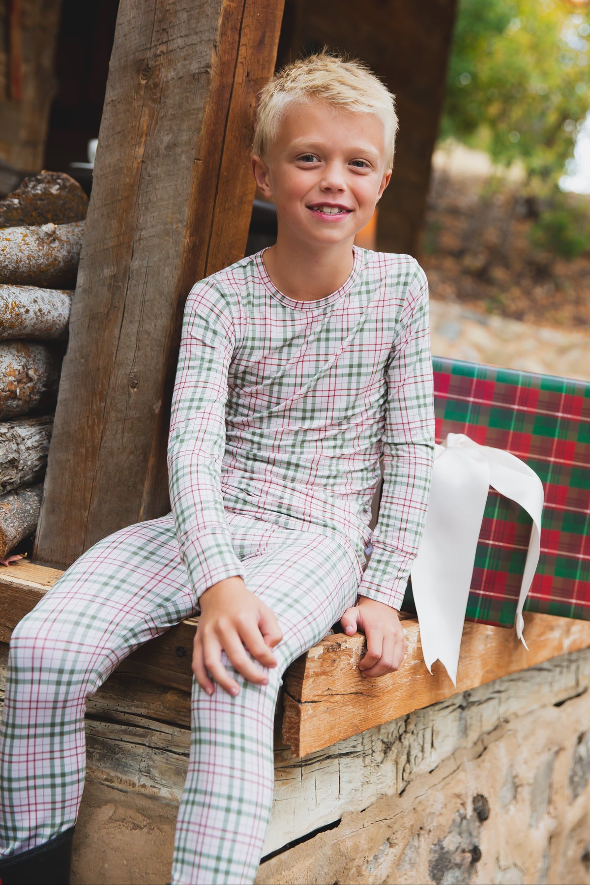 Smiling young boy in homestead / two piece skinny sits on a wooden porch next to a large, wrapped gift with a white ribbon. Logs and trees can be seen in the background.