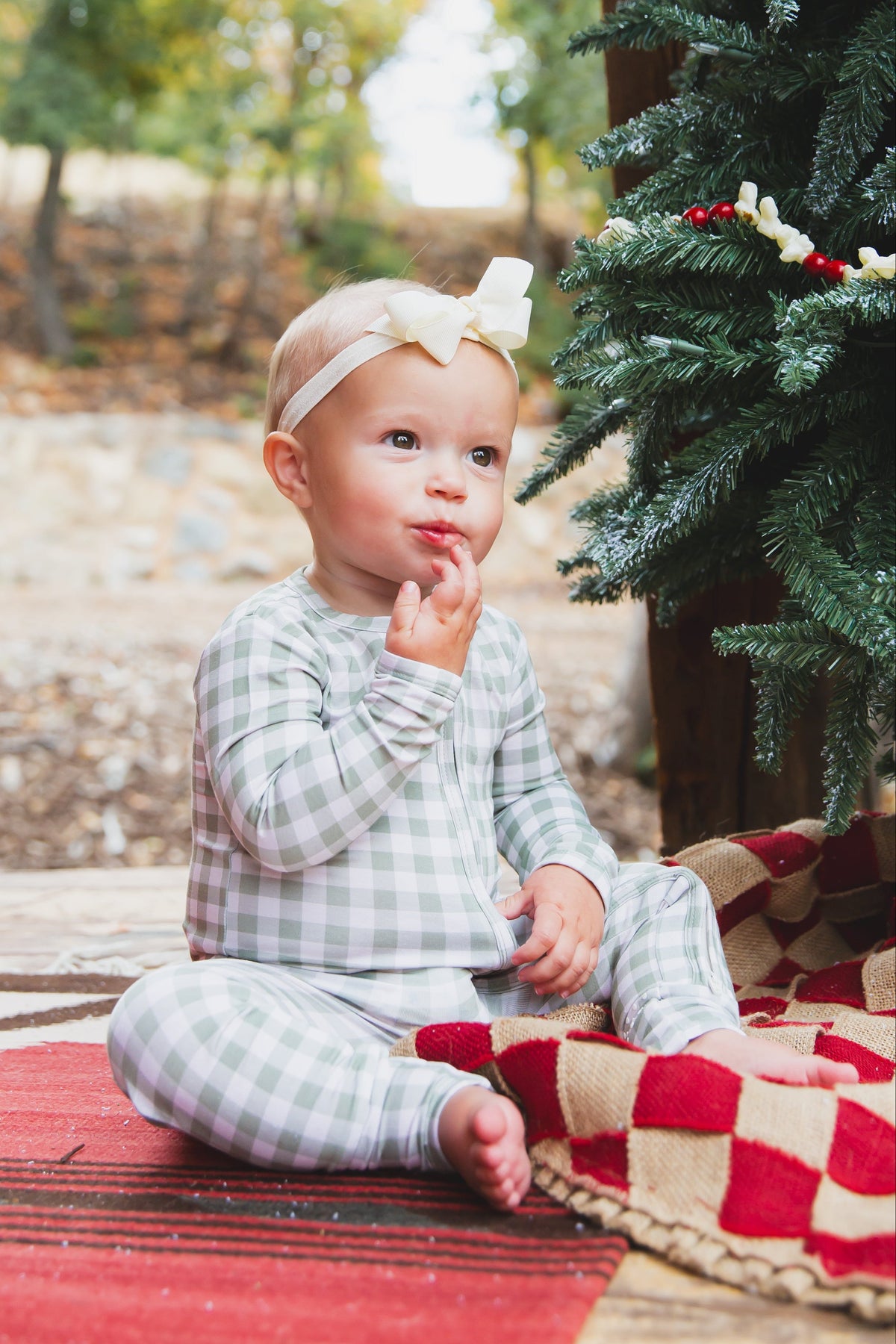 A toddler wearing a green gingham evergreen lane / one piece zipper with a bow headband, sitting on a blanket next to a Christmas tree.