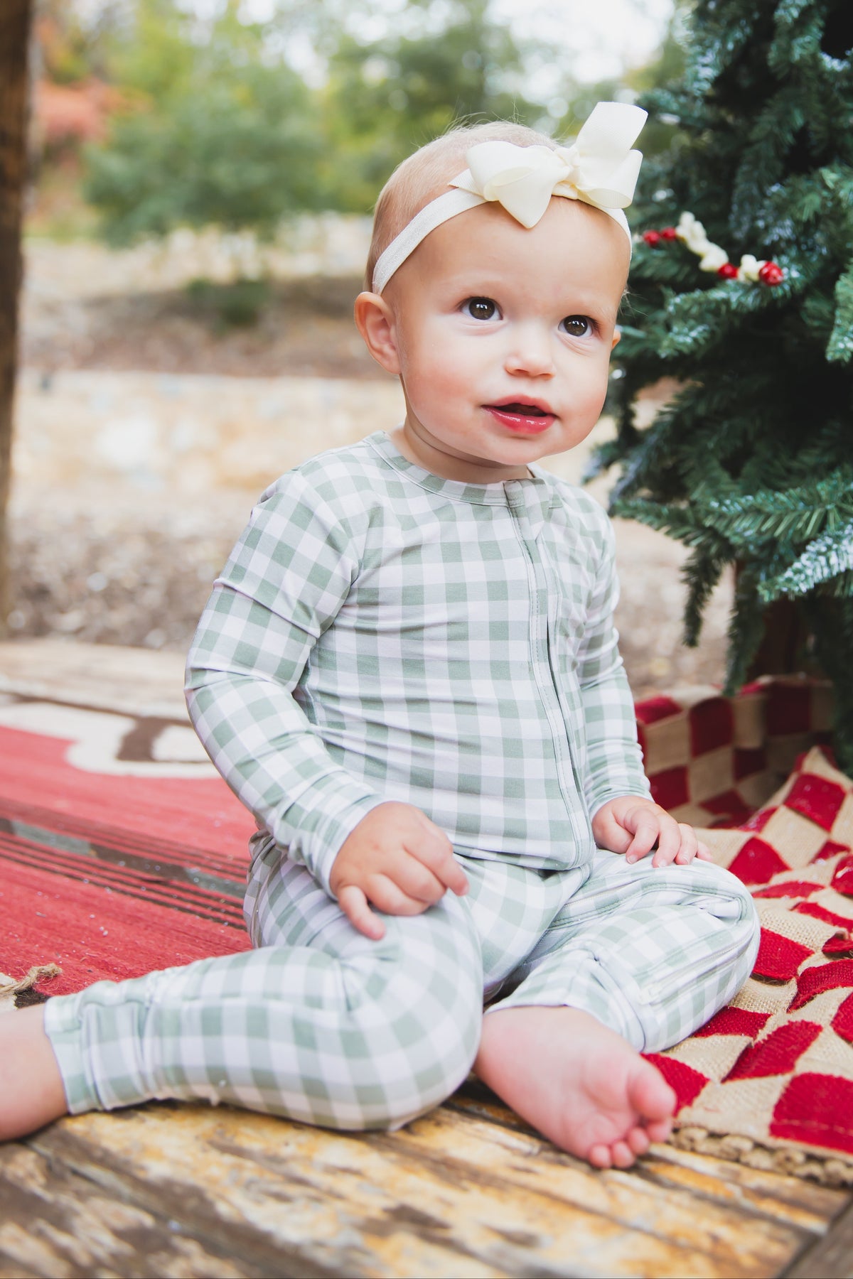 A baby wearing an evergreen lane / one piece zipper in green gingham and a white hair bow, sitting on a red and tan blanket next to a Christmas tree.