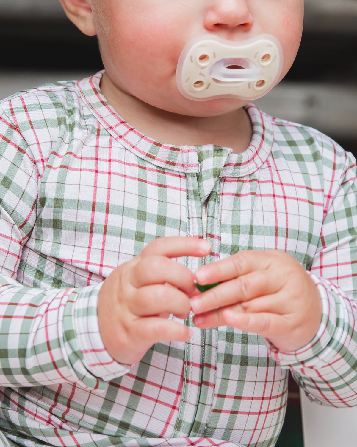 A toddler wearing green and red plaid homestead / one piece zipper pajamas with a pacifier in their mouth.