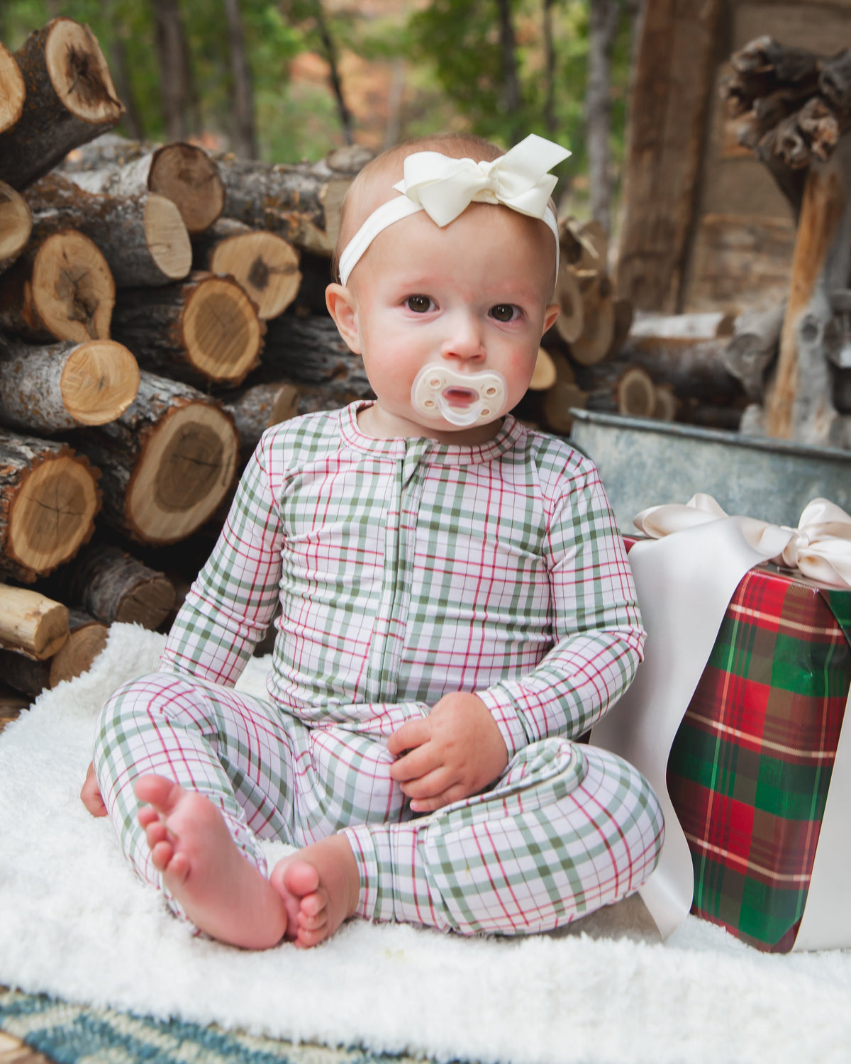 A baby in a long-sleeved plaid homestead / one piece zipper and a white hair bow sits on a white blanket next to a wrapped gift with a large ribbon.