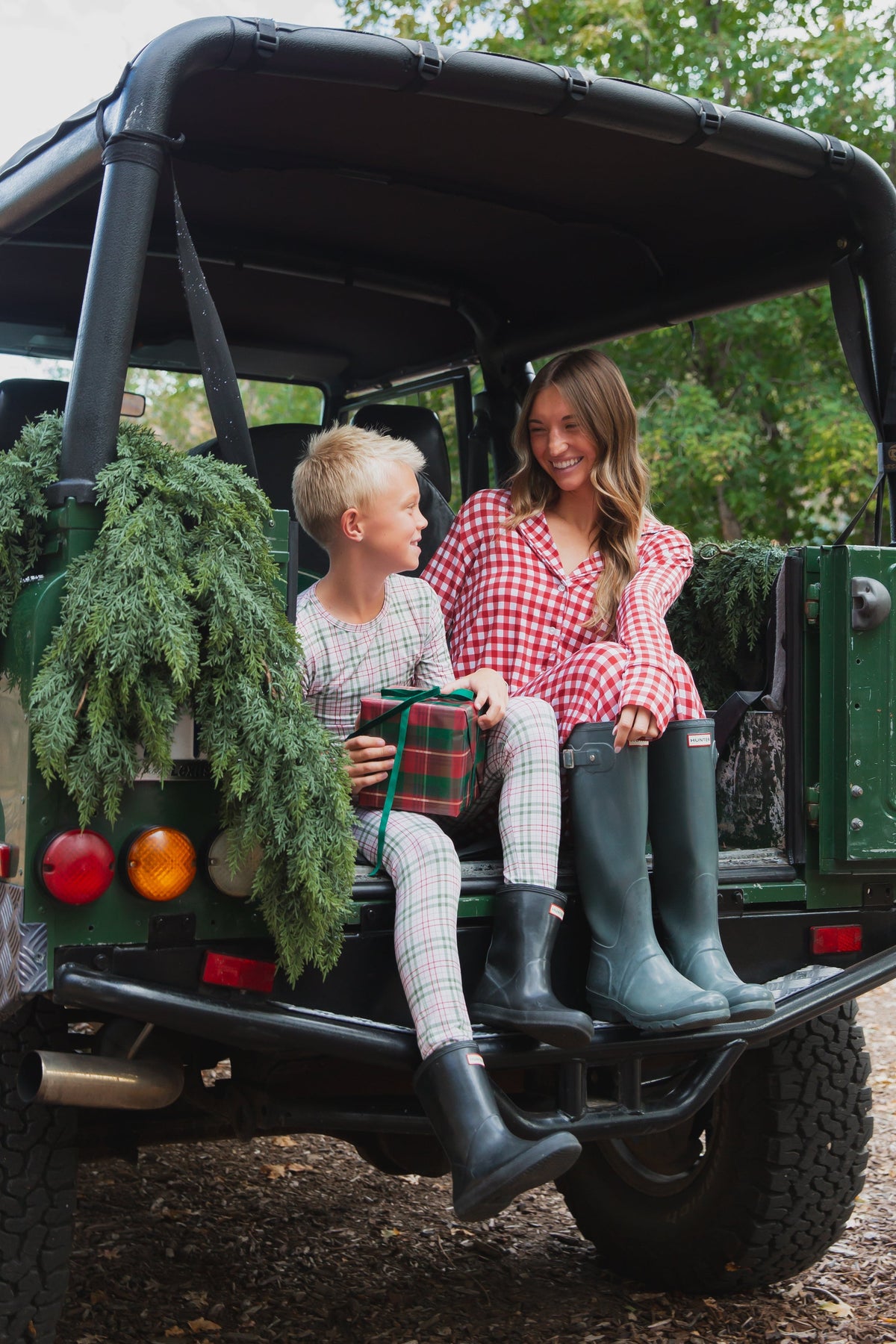 A woman and a young boy wearing homestead / two piece skinny plaid bamboo pajamas and rubber boots sit in the back of an open vehicle holding a gift.