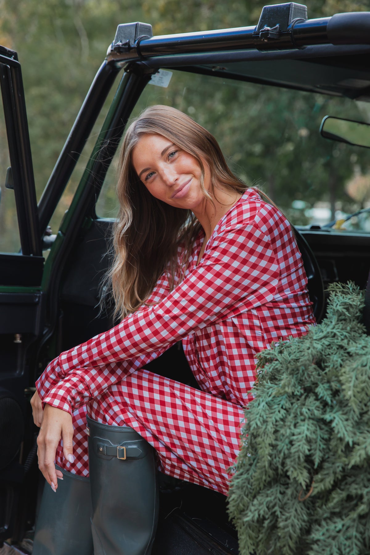 A woman wearing a checkin’ it twice / pajama set / long pant in red and white gingham and dark rubber boots sits in a vehicle next to a small evergreen tree.