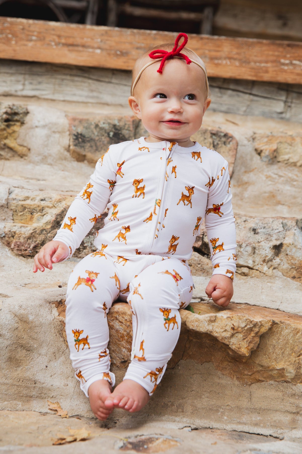 Baby sitting on stone steps wearing a white long-sleeve zippered vintage reindeer / one piece zipper and a red hair bow.