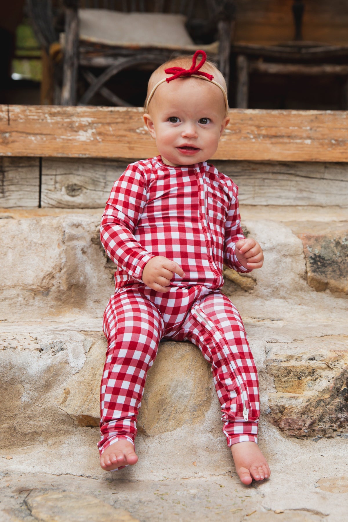 A baby wearing red and white gingham checkin’ it twice / one piece zipper bamboo pajamas sits on stone steps.