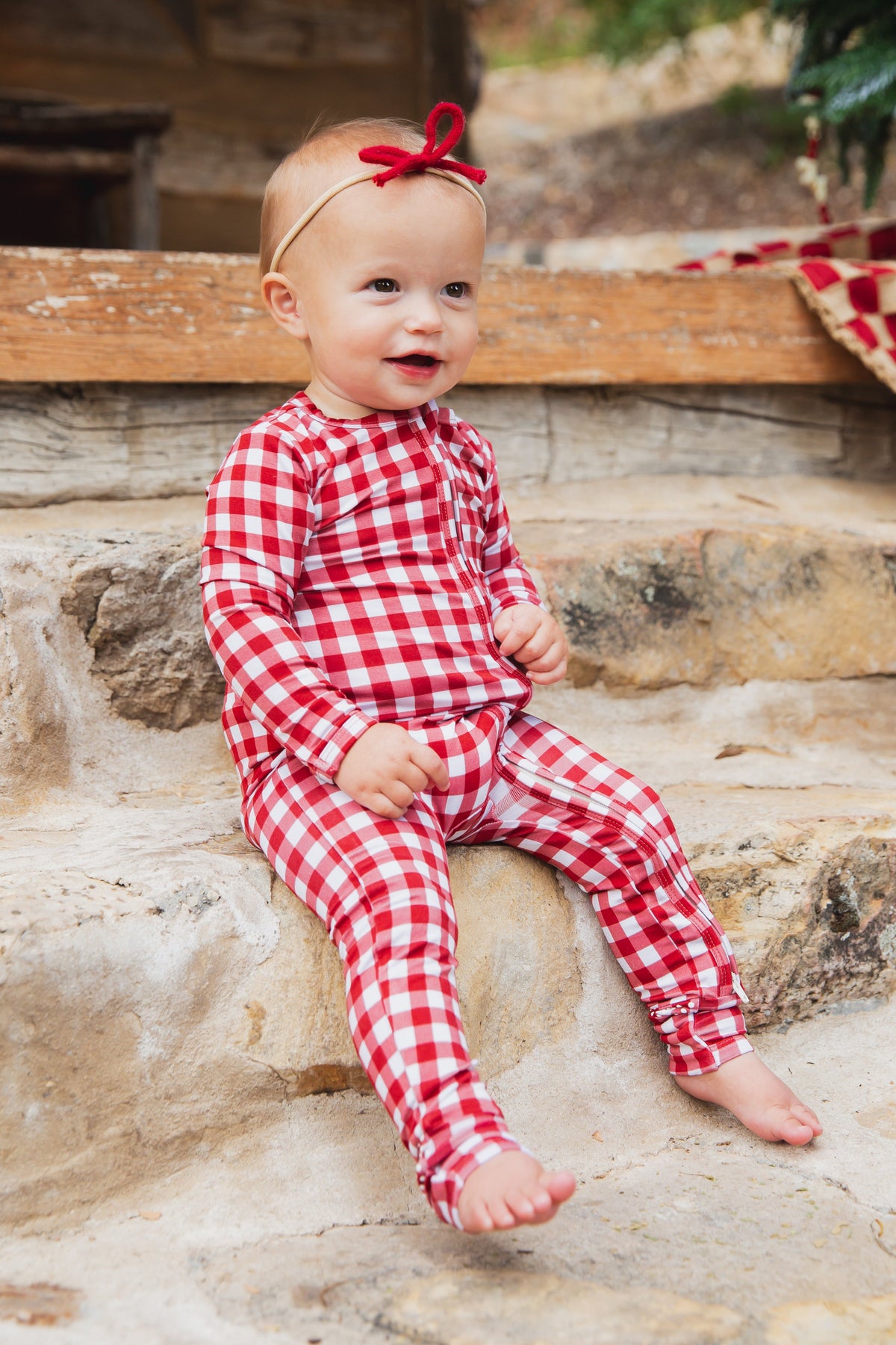 A baby wearing long-sleeved, red and white gingham checkin’ it twice / one piece zipper footie pajamas sitting on stone steps.
