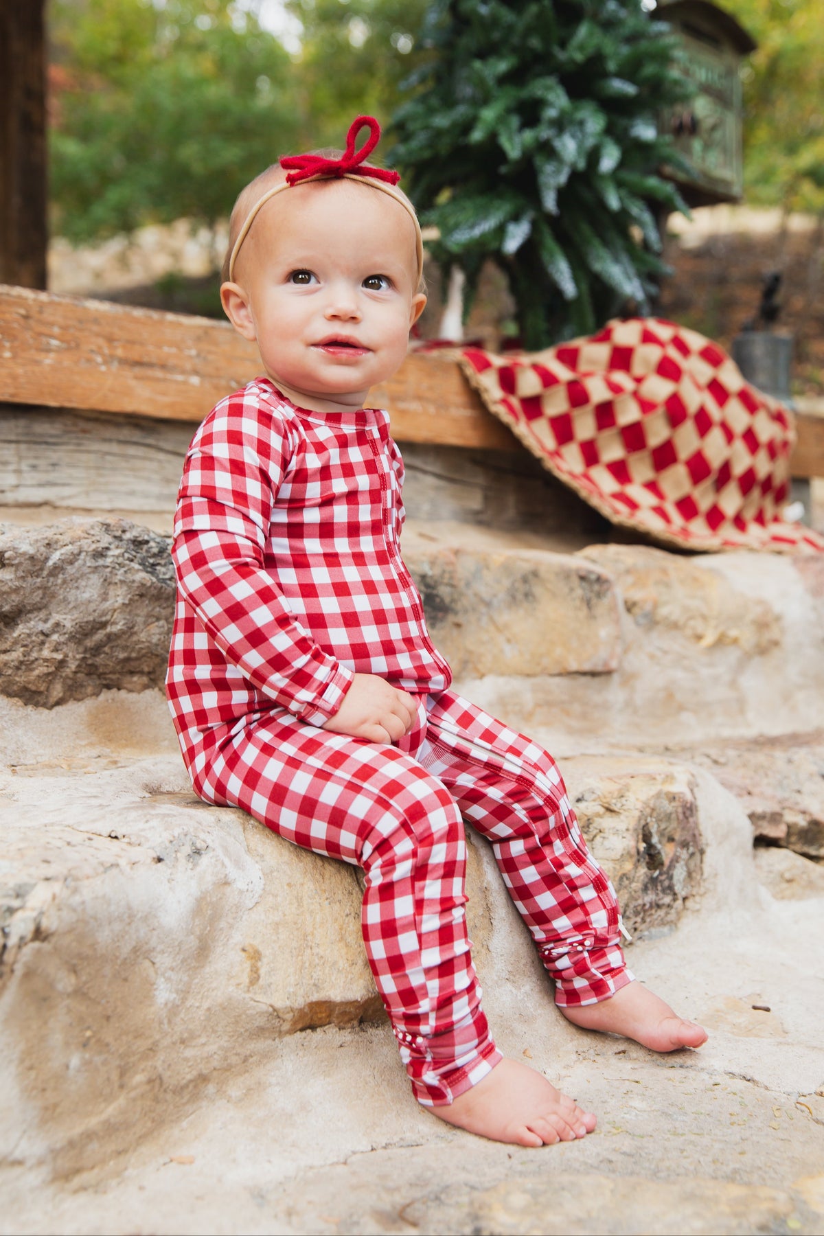 A toddler wearing red and white gingham checkin’ it twice / one piece zipper pajamas and a red bow headband, sitting on stone steps.