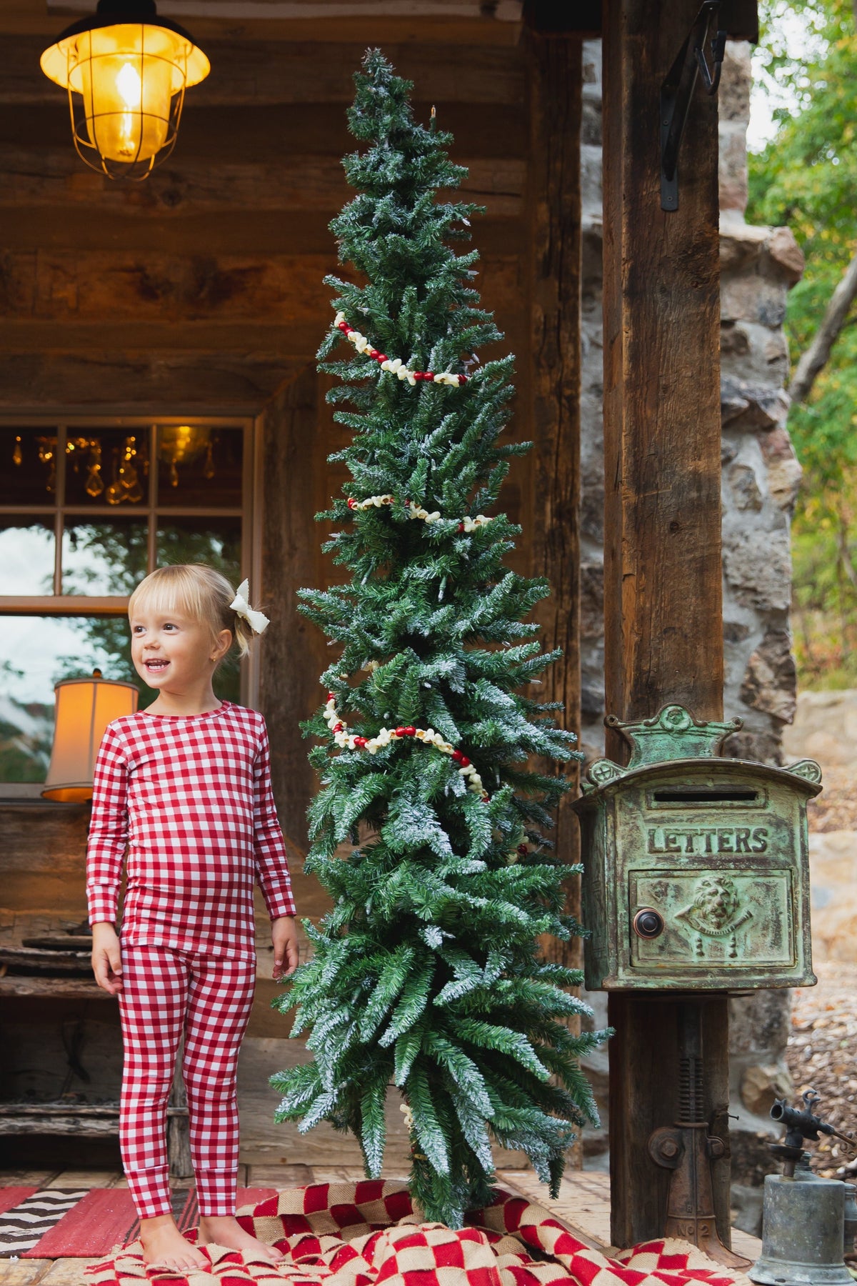A toddler in red and white gingham checkin’ it twice / two piece skinny bamboo pajamas stands next to a slim, flocked Christmas tree on a wooden porch.