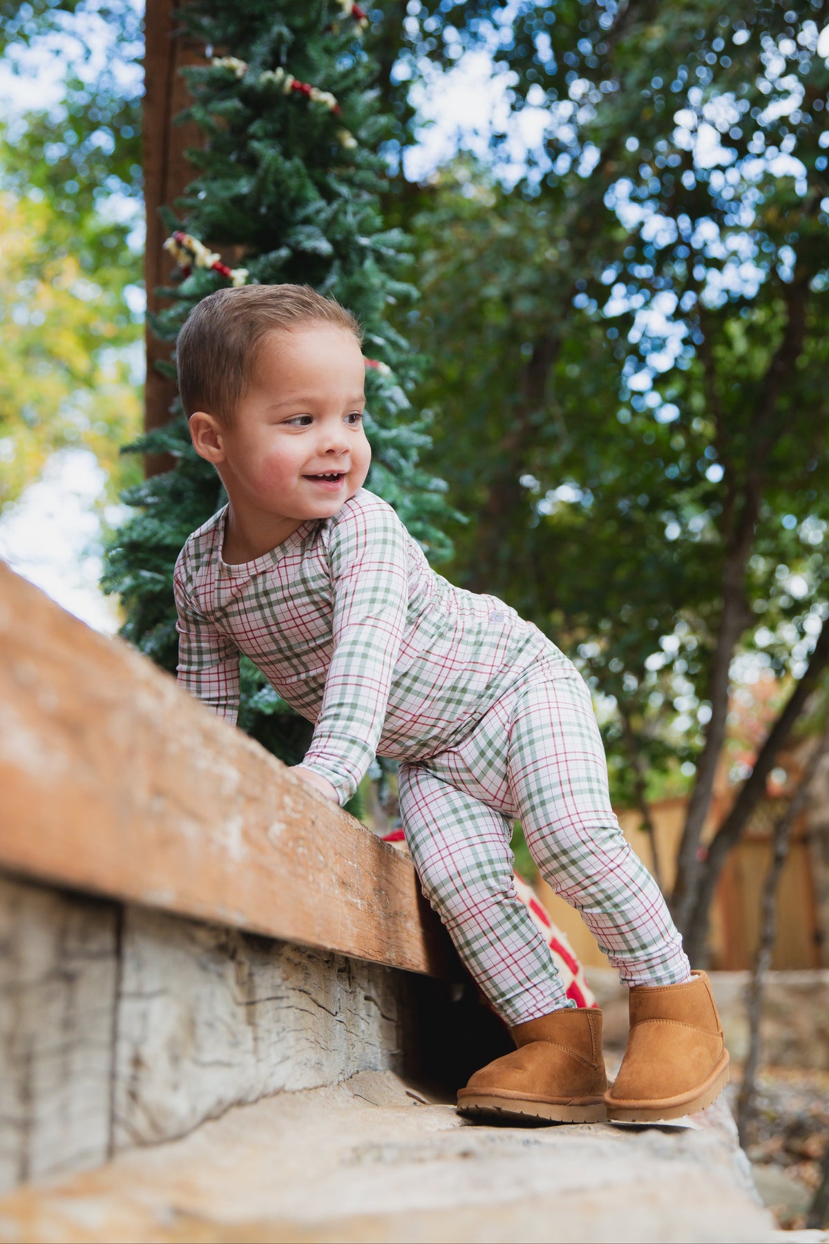 A toddler wearing red, green, and tan plaid homestead / two piece skinny bamboo pajamas consisting of a long-sleeved shirt and leggings with tan ankle boots.
