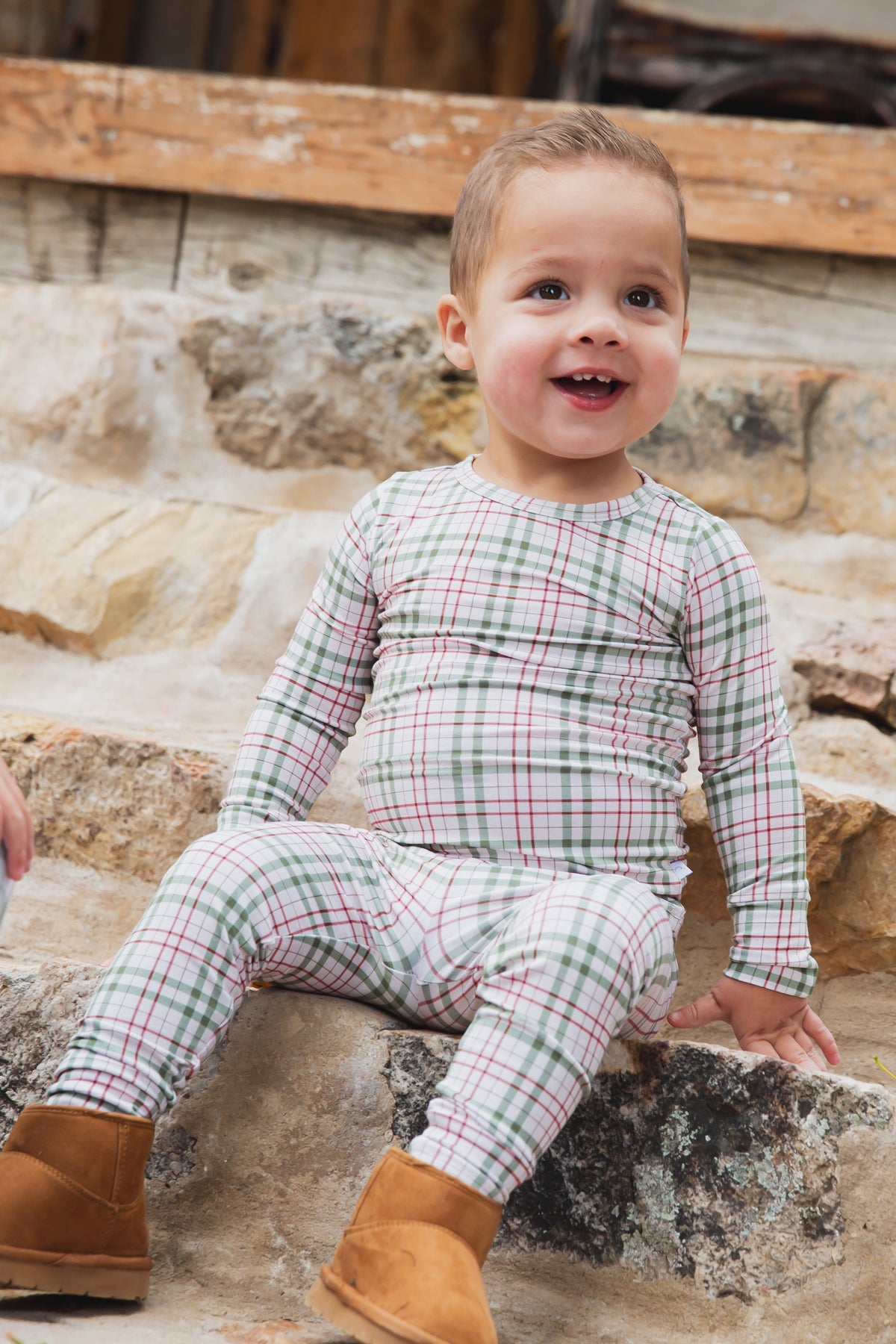 A smiling toddler wearing homestead / two piece skinny green and red plaid pajamas sits on stone steps, paired with brown boots.